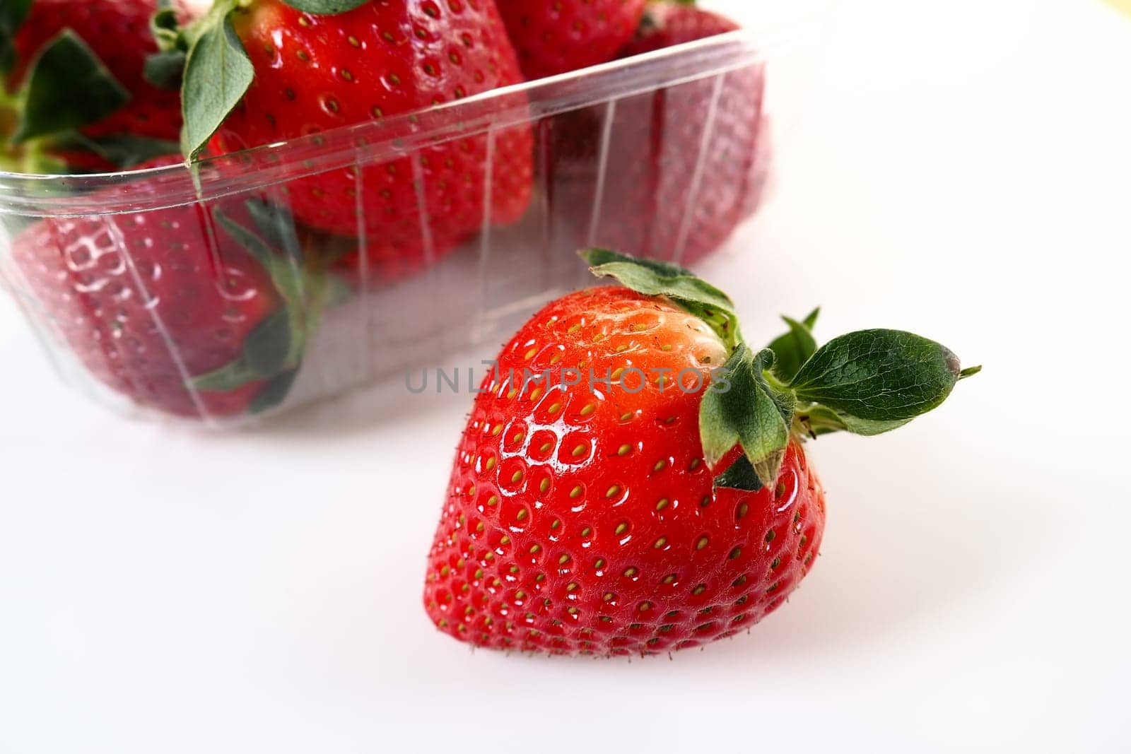 Ripe strawberries in a plastic package on a white background. Delicious fresh berries in a container for sale to customers. Keeping food fresh. Healthy food. One strawberry pulled out of the container
