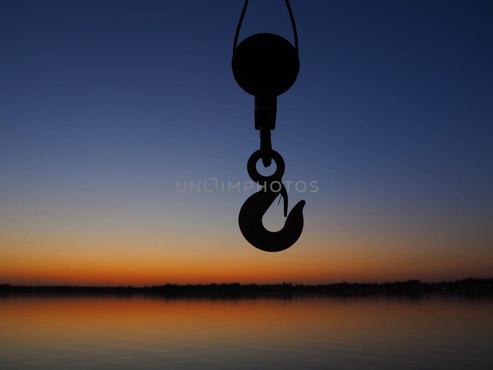 Harbor hook or bracket for transporting goods in a port or pier. The hook is a clue against the horizon, water and sunset. Evening landscape with a hanging device in the foreground.