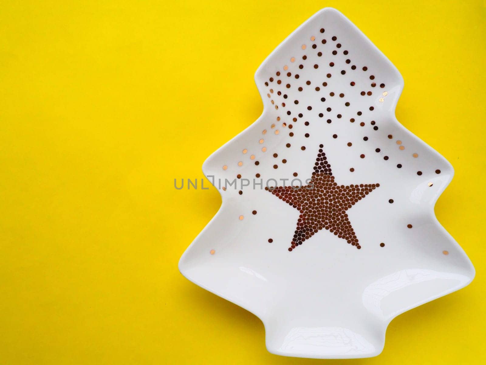 White plate in the shape of a Christmas tree with gold decorations and a star in the middle. Yellow background. New Year or Christmas card. Background for advertising and congratulations. Copy space.