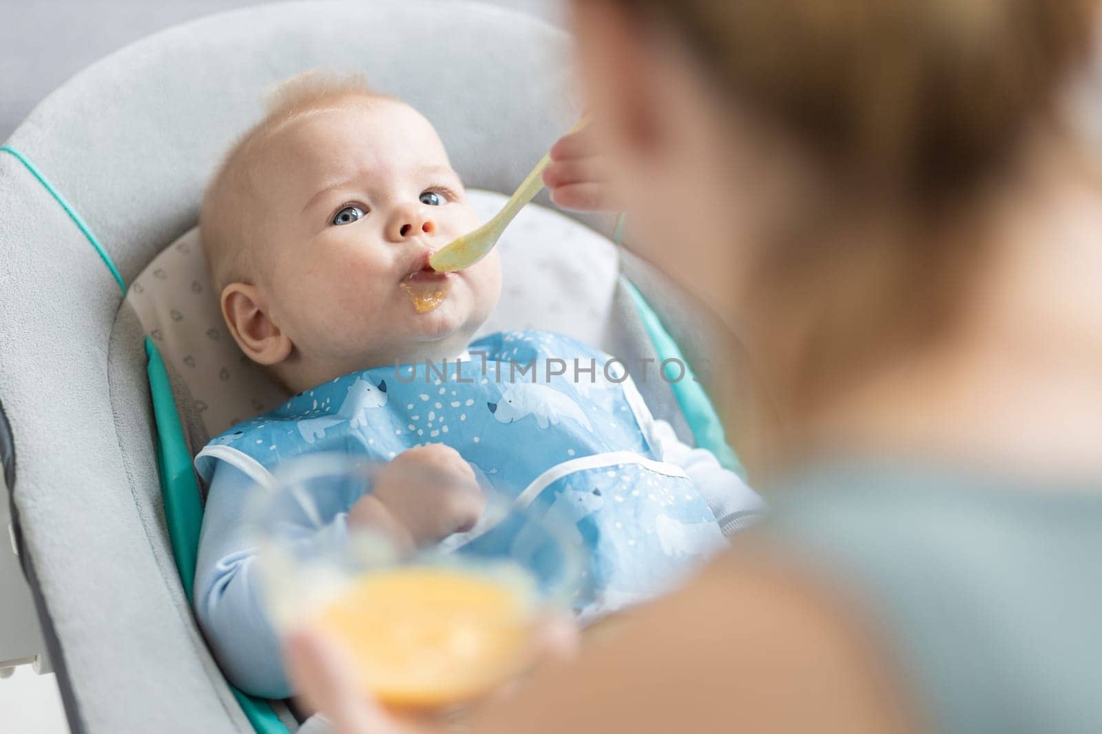 Mother spoon feeding her baby boy child in baby chair with fruit puree. Baby solid food introduction concept