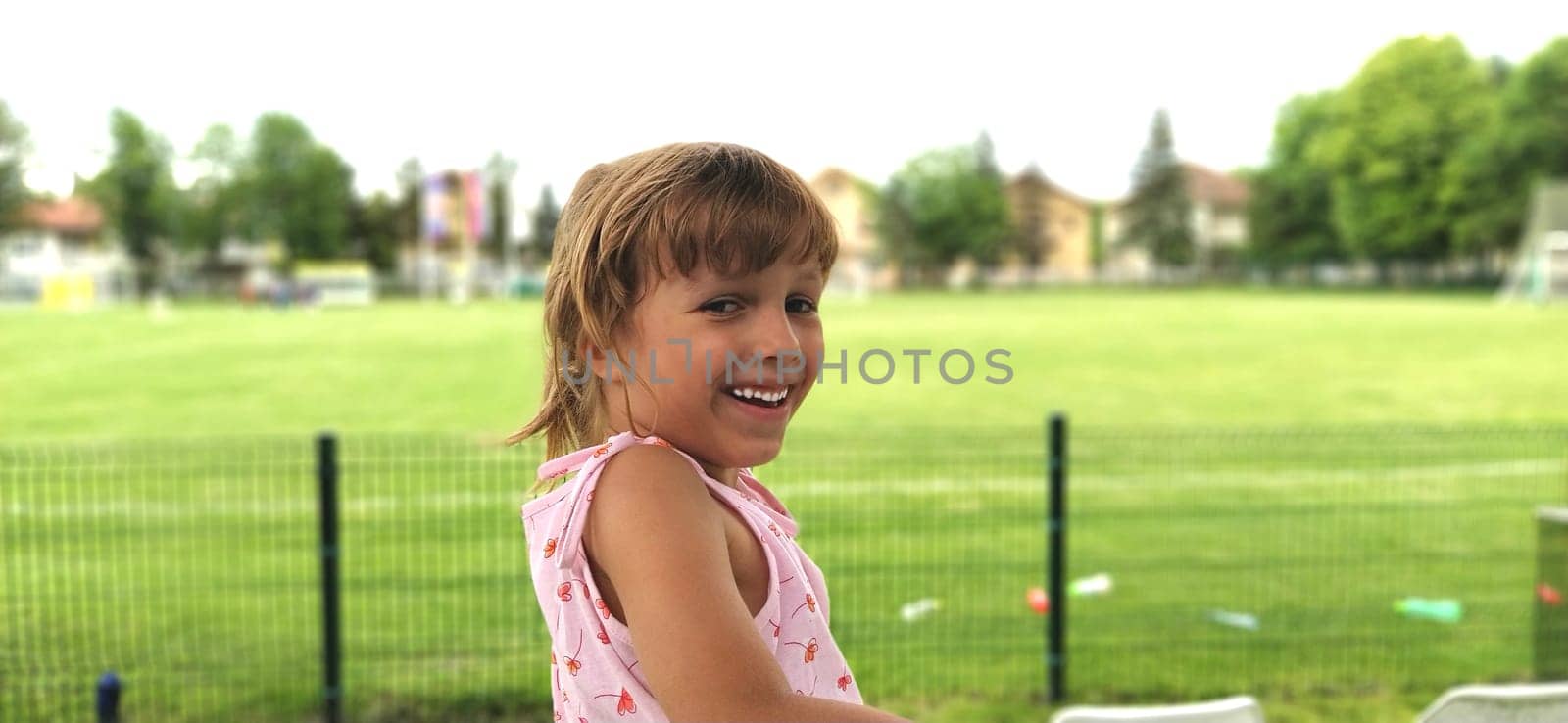 young beautiful tanned girl turns back and smiles. A child on the background of a soccer field. Partially defocused. Baby smile with white teeth. Green grass as a background.