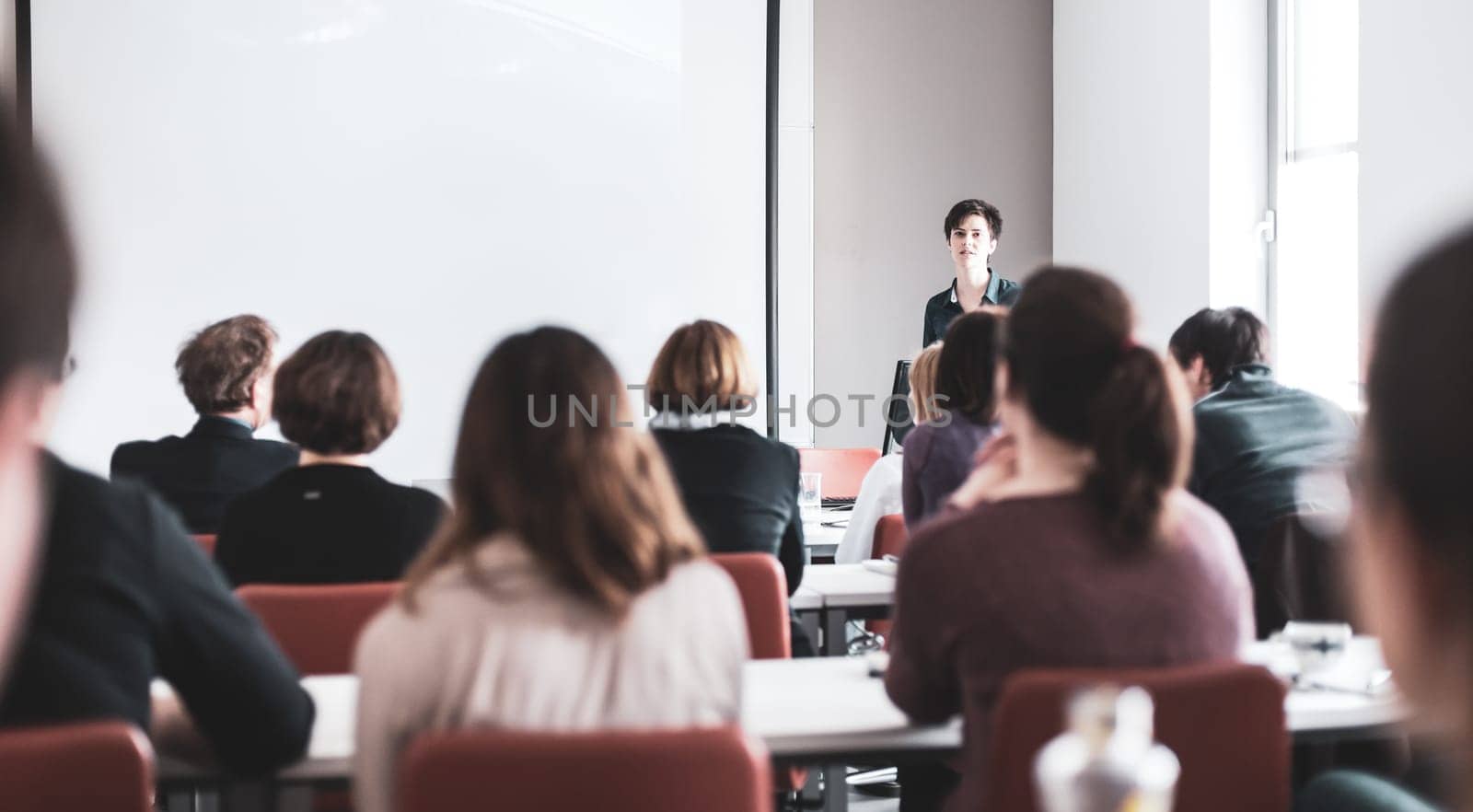 Female speaker giving presentation in lecture hall at university workshop . Participants listening to lecture and making notes. Scientific conference event.