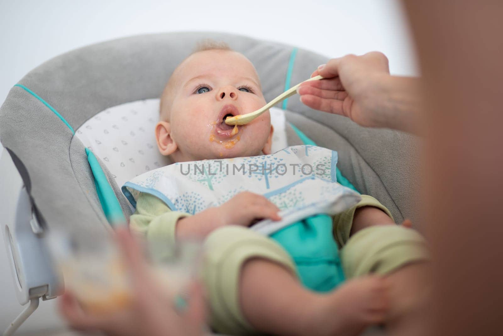 Mother spoon feeding her baby boy child in baby chair with fruit puree. Baby solid food introduction concept
