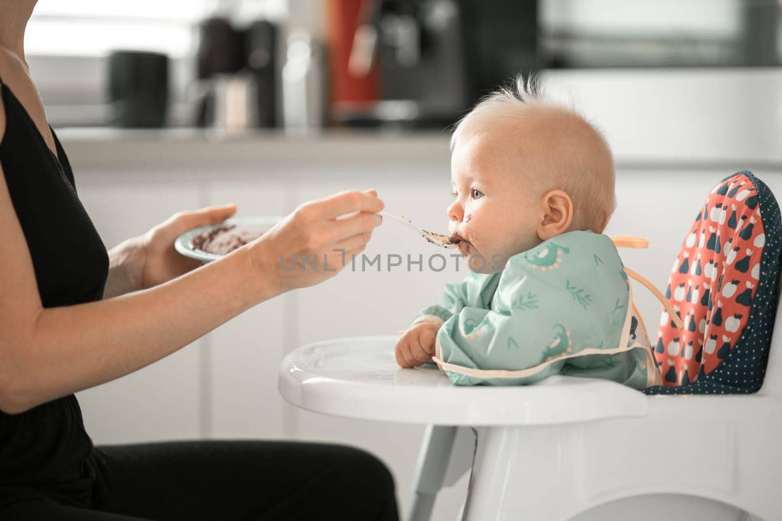 Mother spoon feeding her baby boy child in baby chair with fruit puree in kitchen at home. Baby solid food introduction concept