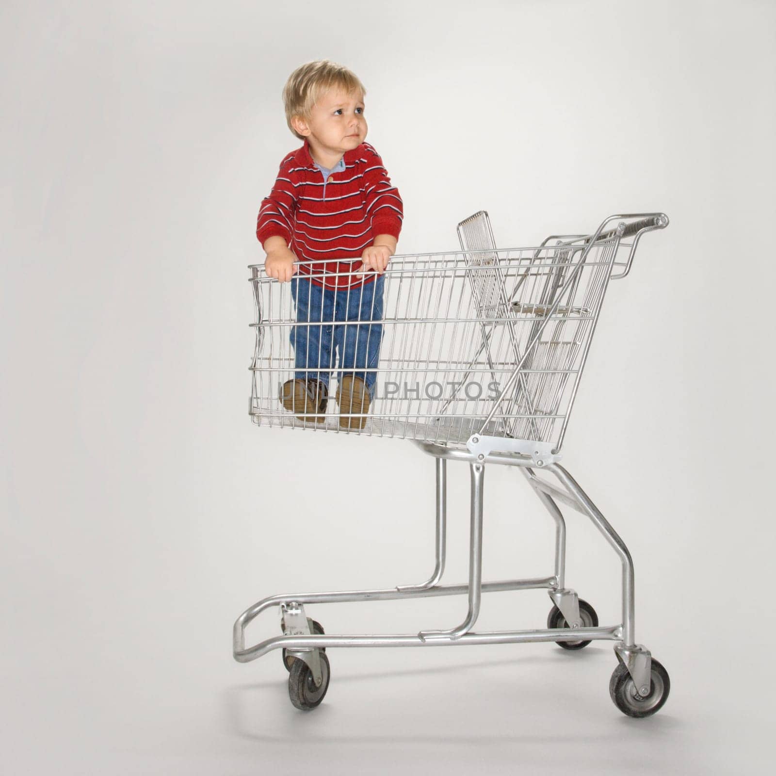 Studio portrait of Caucasian boy standing alone in empty shopping cart against white background.