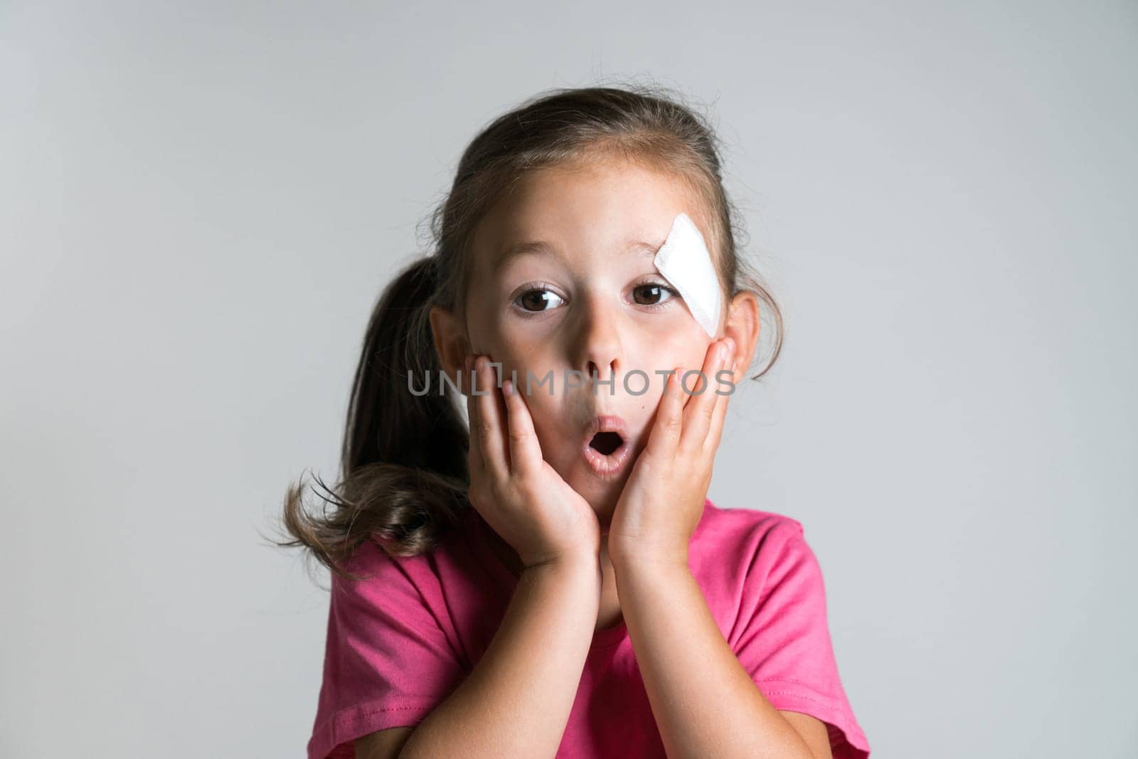 Close up surprised, cute, nice dark haired little girl with bandage patch on face, wear pink shirt on white background. Afraid of parents reaction. Physical wounds, fidget children, doctor fear