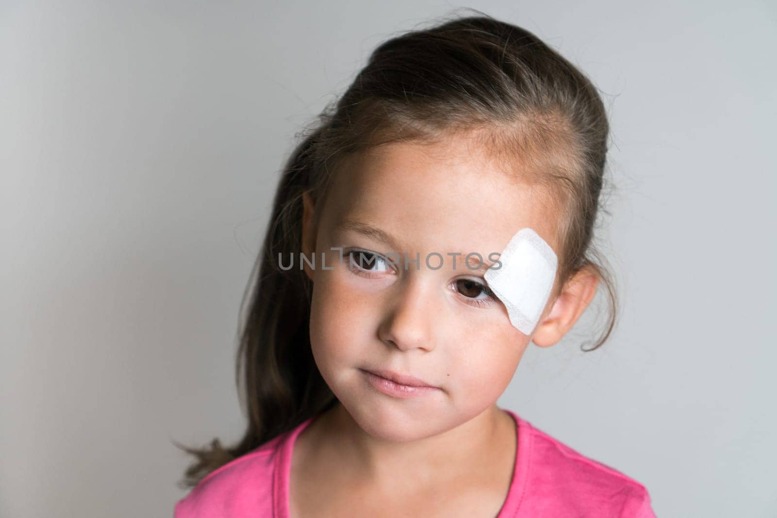Close up sad, nice, lovely, adorable dark haired little child, girl with white patch bandage on face, looking down. Child abuse and physical wounds. Kid protection and safety, healthcare and medicine