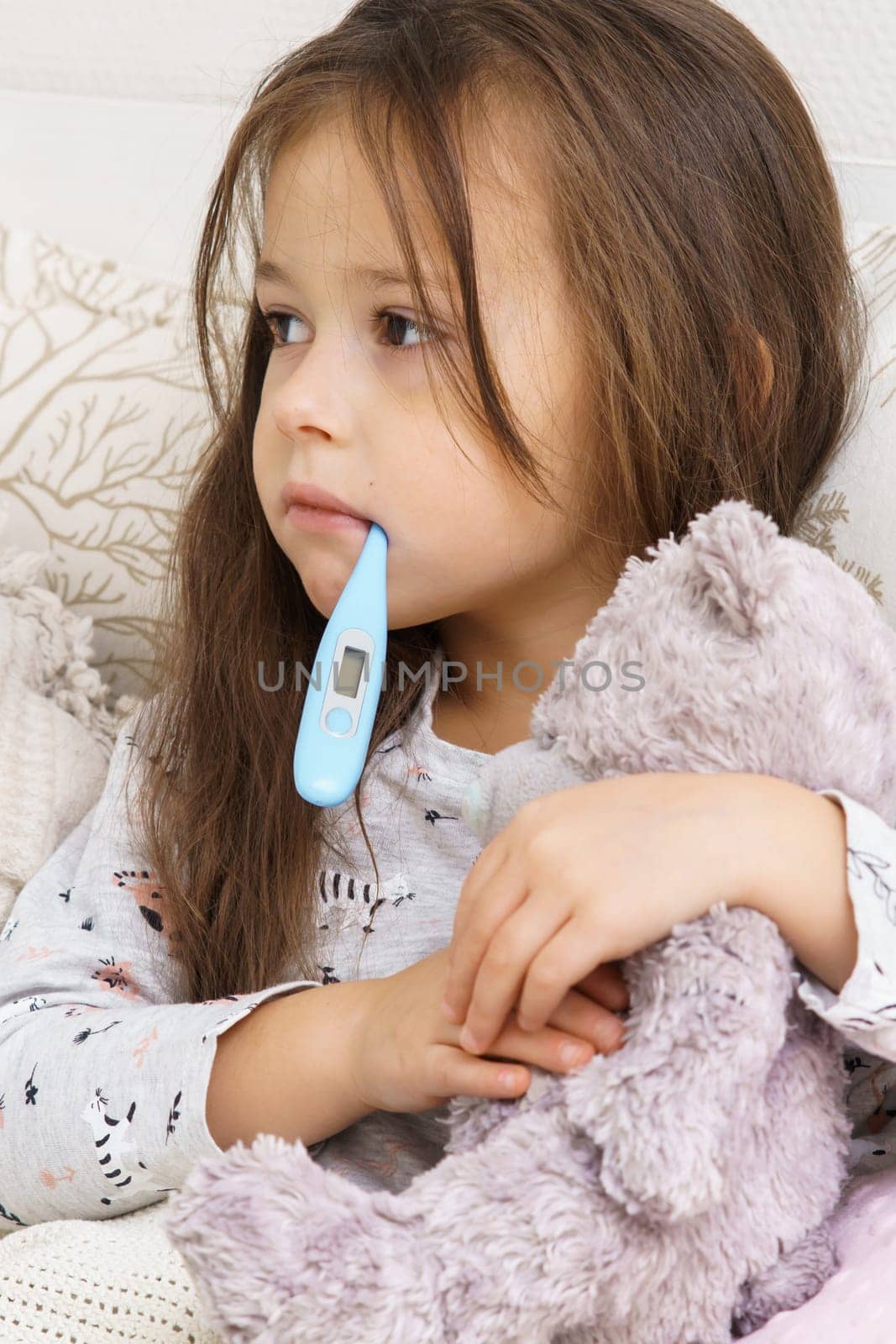 Vertical sad, calm. depressed, tired little child, girl looking away and holding electronic thermometer in mouth, embracing teddy bear in bed. Seasonal cold, influenza and virus infection disease