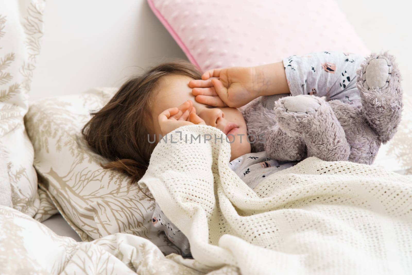 Sad, disappointed, crying child girl rubbing eyes, holding stuffed toy, lying in soft white bed with pillows in the morning. Hurt and family child abuse, hard relationship. Social issues, loneliness