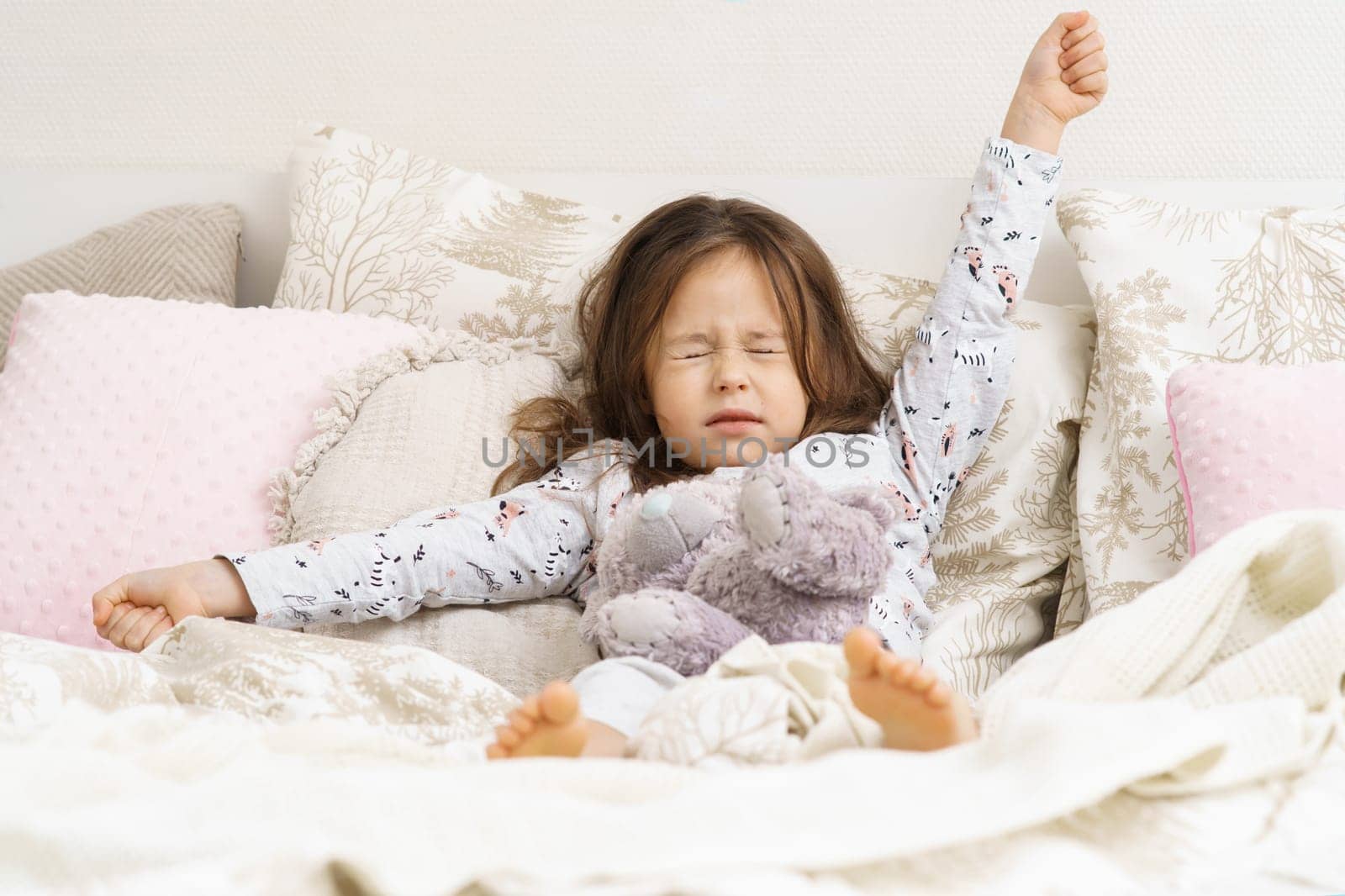 Small adorable, dreamy, cute kid, girl stretching oneself with raising arms, close eyes, with stuffed bear toy in soft white bed in the morning. Lazy morning, waking up and children sleepless