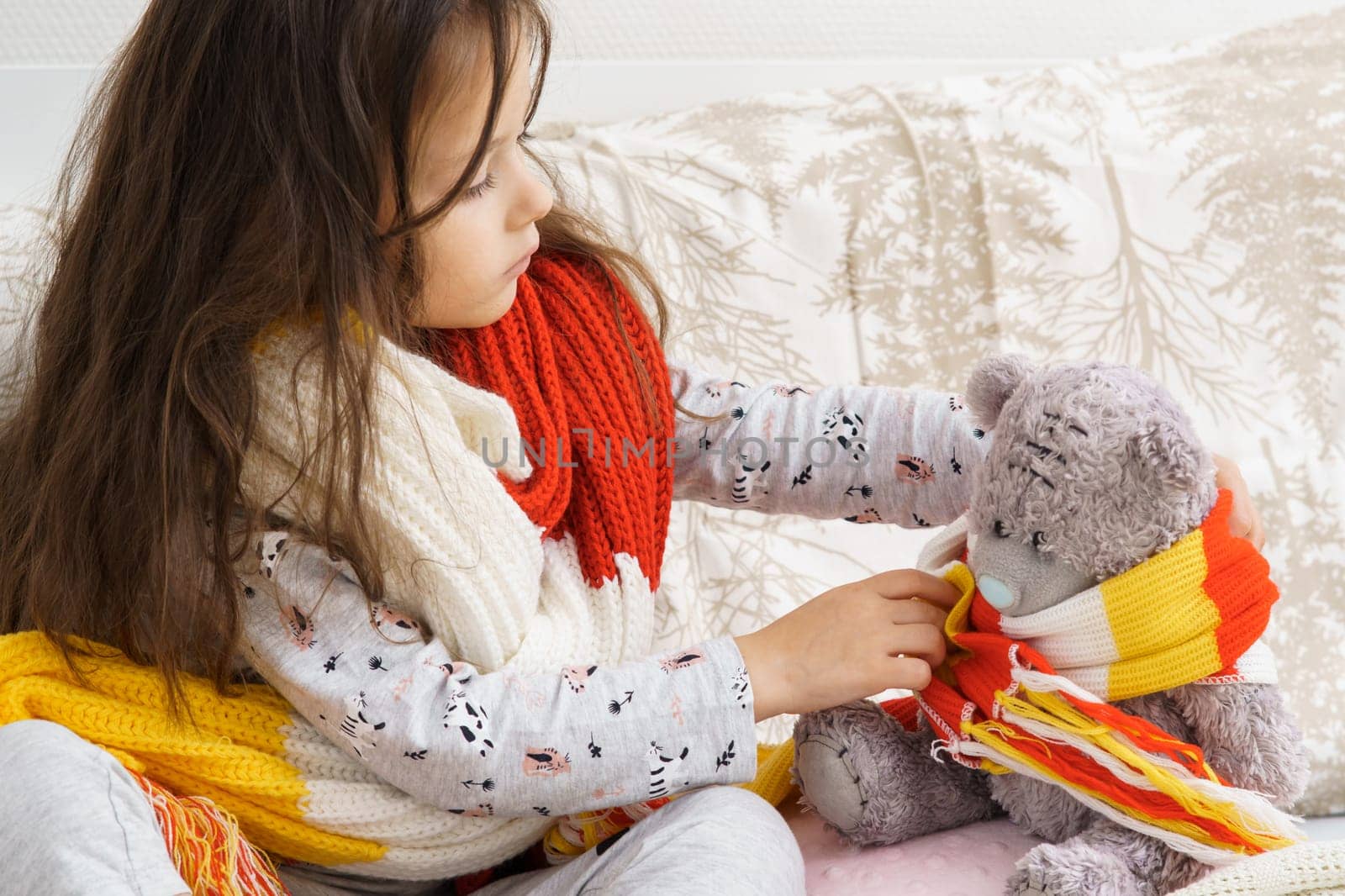 Serious child, girl healing bear toy friend, wrapping in red, yellow scarf, sit on bed in bedroom. Little doctor help best friend. Illness, sickness and seasonal flue. Sore throat, angina symptoms