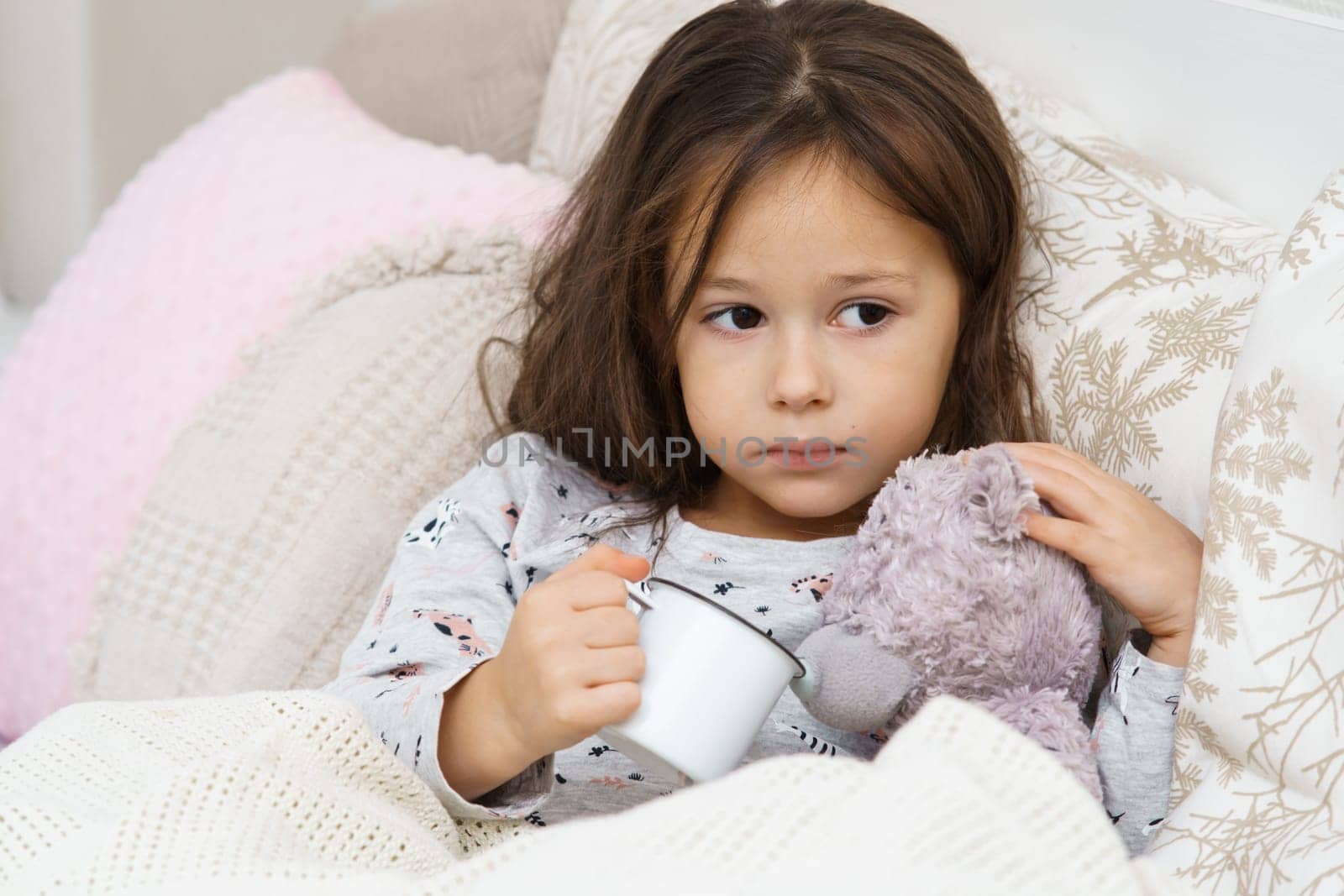 Sad, worried, calm little dark haired preschooler girl watering bear toy from mug, lying in pajamas in soft white bed. Recovering, health care. Natural vitamin tea or liquid medicine. Bed resting