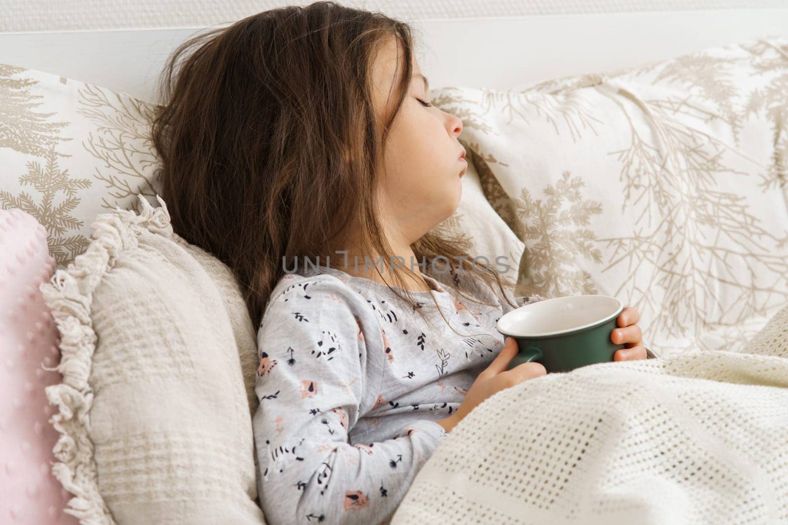 Dear little dark haired preschooler girl coughing, drinking hot tea or medicine from green mug, lying in pajamas in soft bed. Sore throat, angina, tonsillitis. Recovering, health care, home treatment