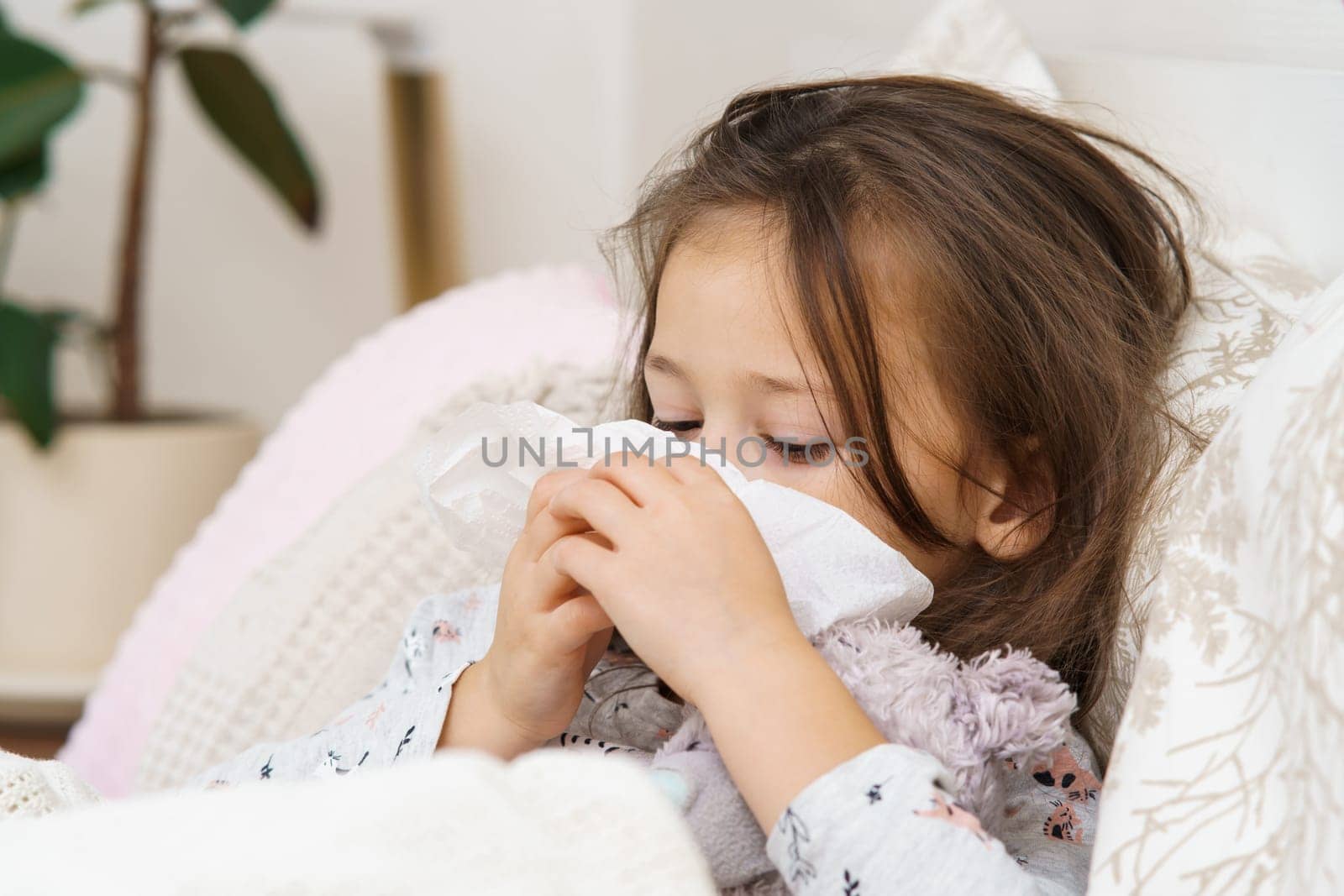 Sad, depressed little long haired girl covering face with napkin tissues, lying with soft toy, wrapped in blanket. Flu season, coronavirus and allergy sypthoms with seasonal infection. Bedtime at home