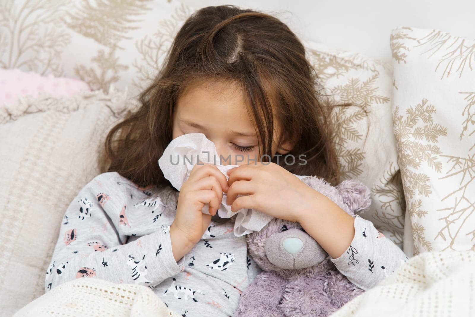 Tired, sleepy little brunette girl sneezing snot with tissues, embracing bear toy in soft bed. Headache and rhinitis symptoms with seasonal viral infection. Bedtime at home, lockdown and quarantine