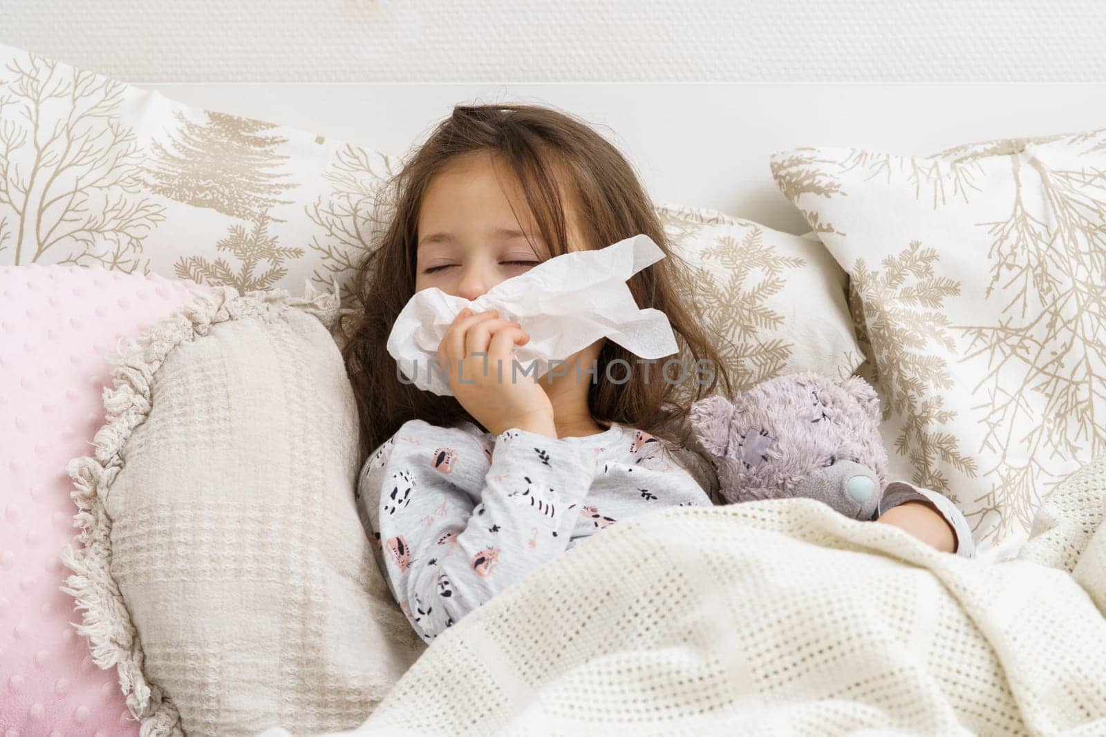 Sleepy, exhausted little brunette girl with close eyes wiping wet nose with tissues, lying with bear toy in bed. Flu season and rhinitis, headache and allergy with seasonal infection. Fever and cold
