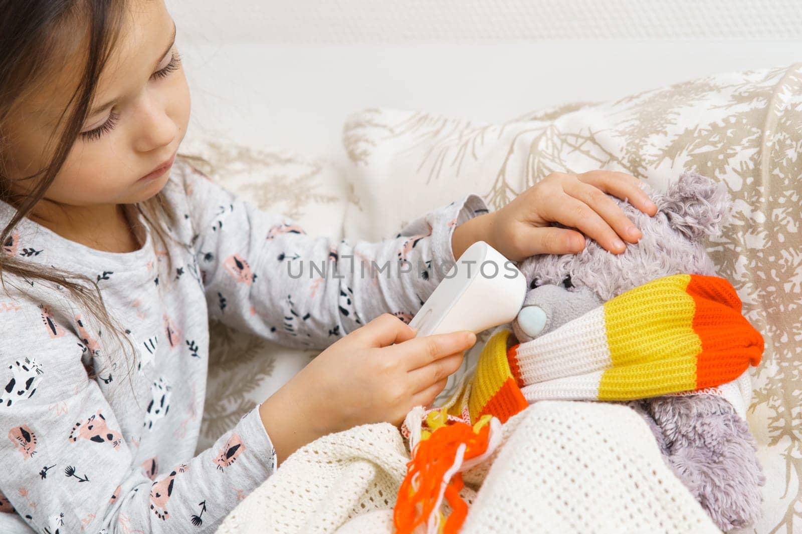 Concentrated child, girl healing bear toy, examining temperature with electronic thermometer, wrapping in scarf on bed, sit on sofa. Illness, sickness and seasonal flue. Little doctor help best friend