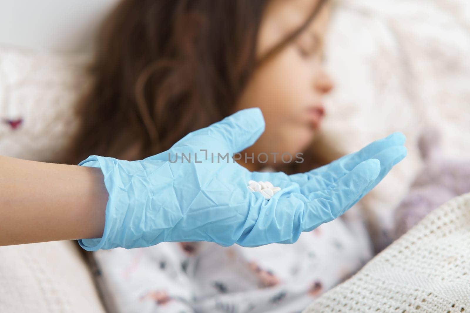 Cropped doctor hand in blue sterile glove holding white pills against defocused unhealthy little brunette girl sleeping in bed. Healthcare and pharmacy recovery. Sickness, illness and virus infection.