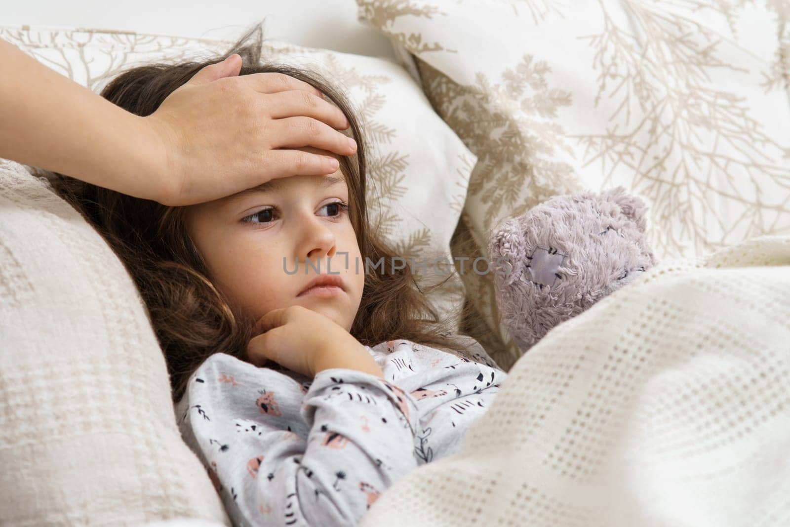 Unrecognizable woman hand touching little girl forehead in bedroom. Sad, indifferent, bored little brunette girl lying with bear toy in white soft bed. Pediatrician healthcare and protection