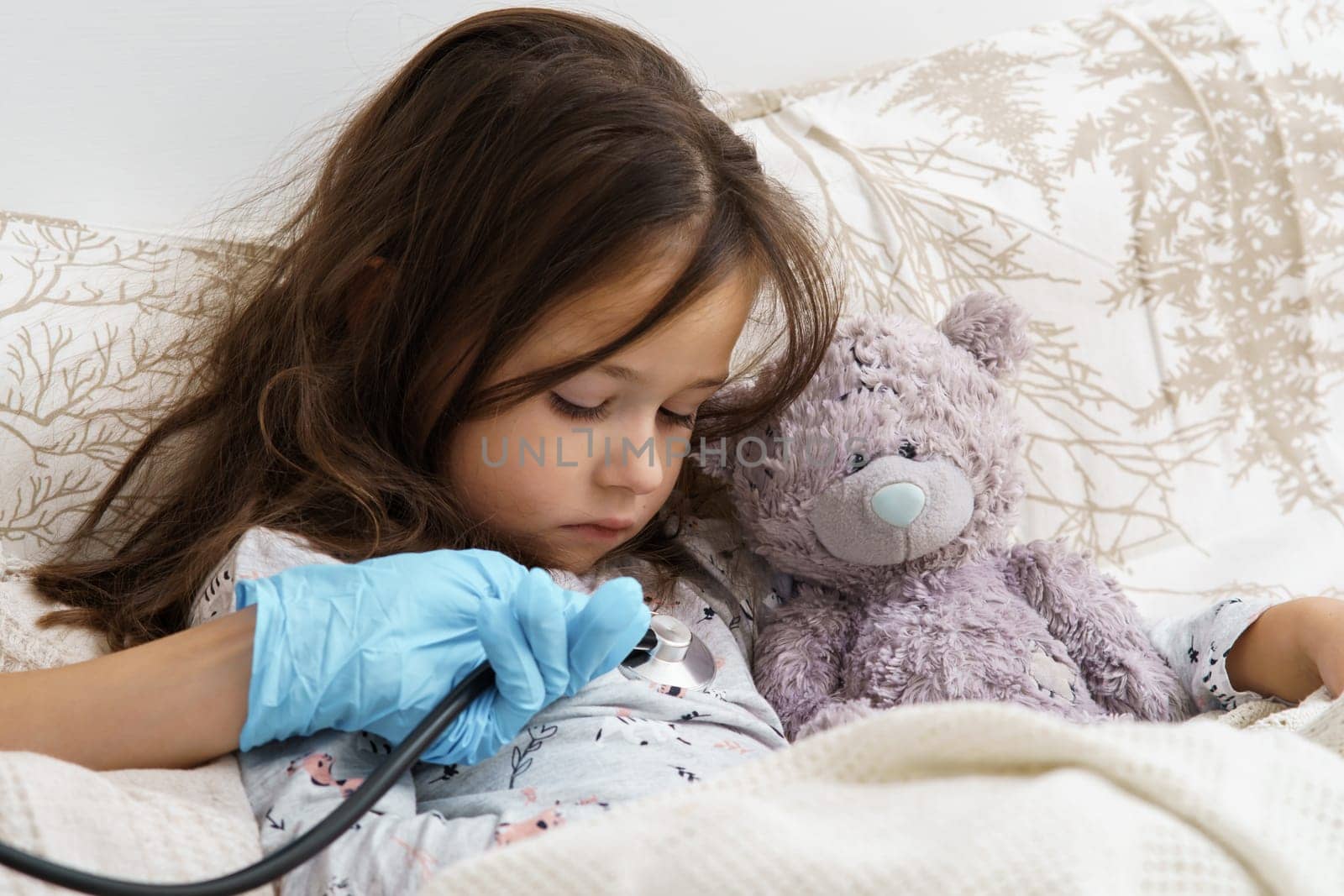 Cropped doctor hand in sterile glove using stethoscope to listen to heart and lungs of little girl lying in bed with bear toy. Seasonal infection, virus, coronavirus. Healthcare and medicine