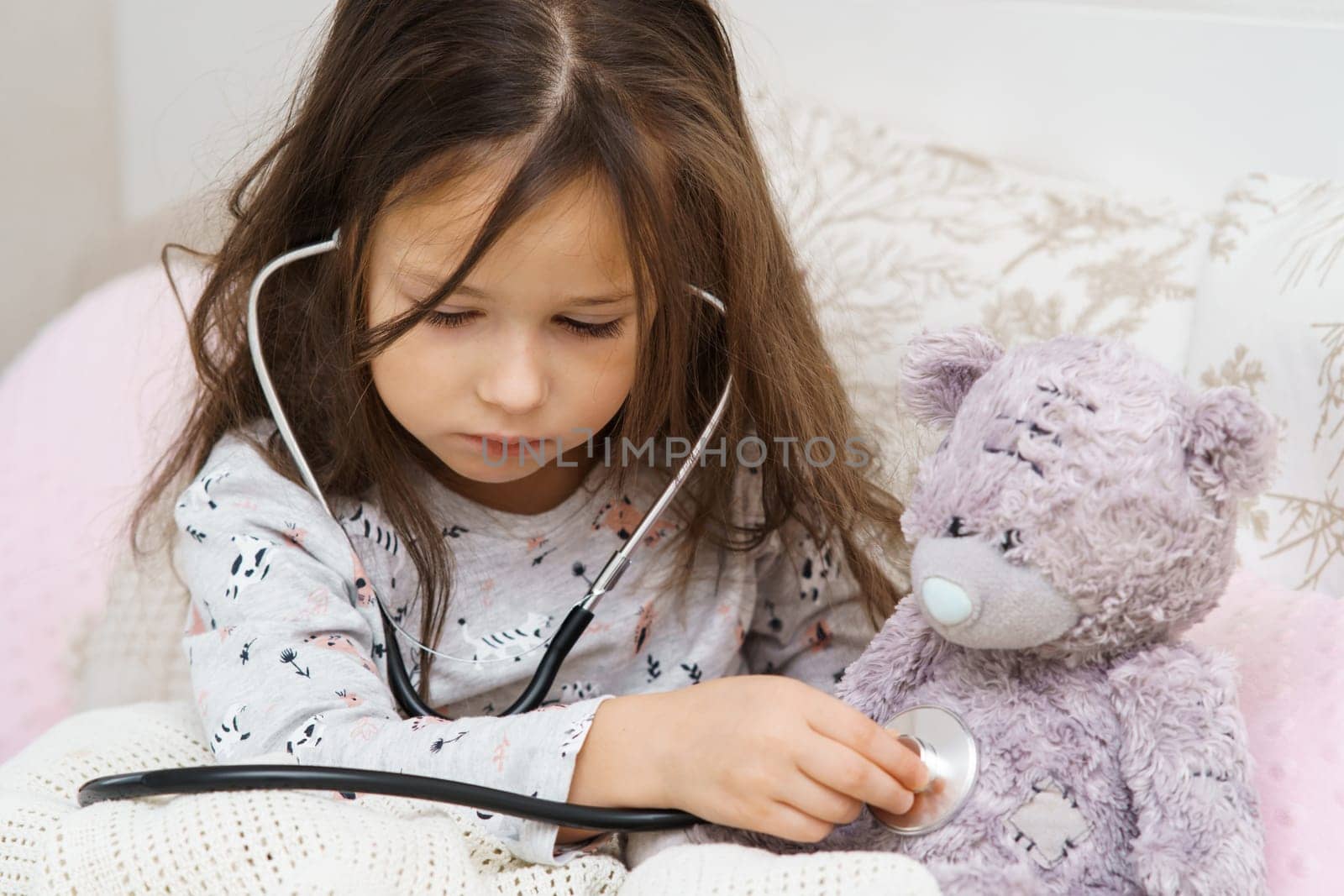 Serious, concentrated, careful small child, girl checking teddy bear chest, breathing with stethoscope, wrapped in blanket. Healthcare and medicine. Little doctor help at home. Friends sickness