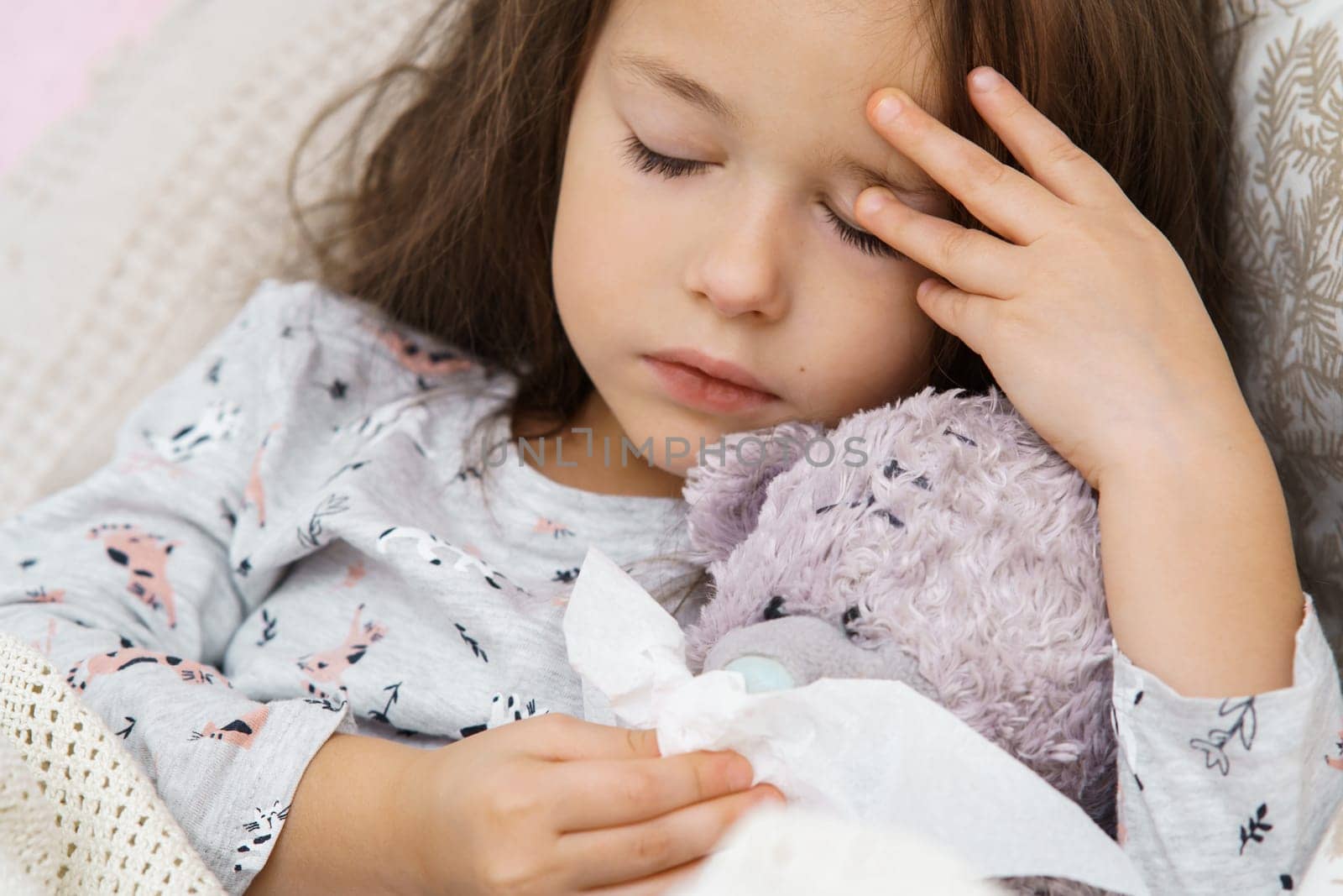Tired little brunette girl napping with close eyes, touching head, wiping imaginary snot of bear toy with tissues. Fever and cold. Flu season ahead, headache and allergy with seasonal infection.