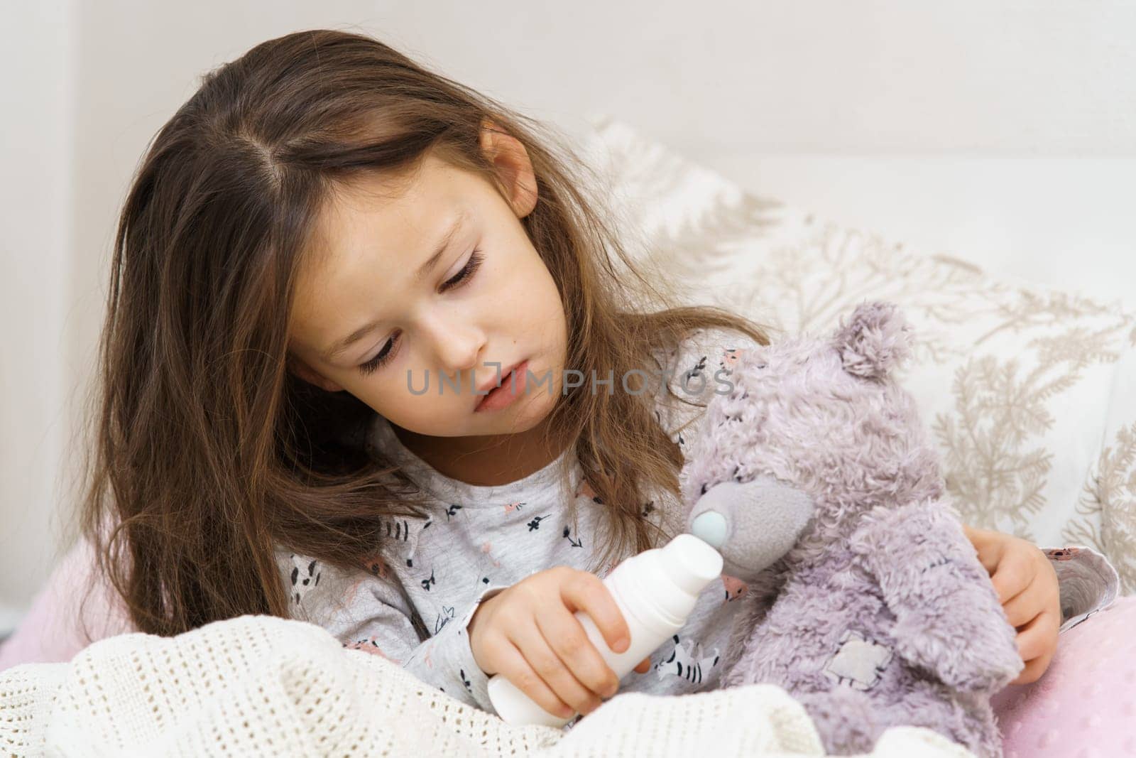 Worried, sunless, tired little brunette girl embracing bear toy patient, healing with nose spray, sit on bed in bedroom. Lockdown isolation, loneliness at home. Children sickness, little doctor