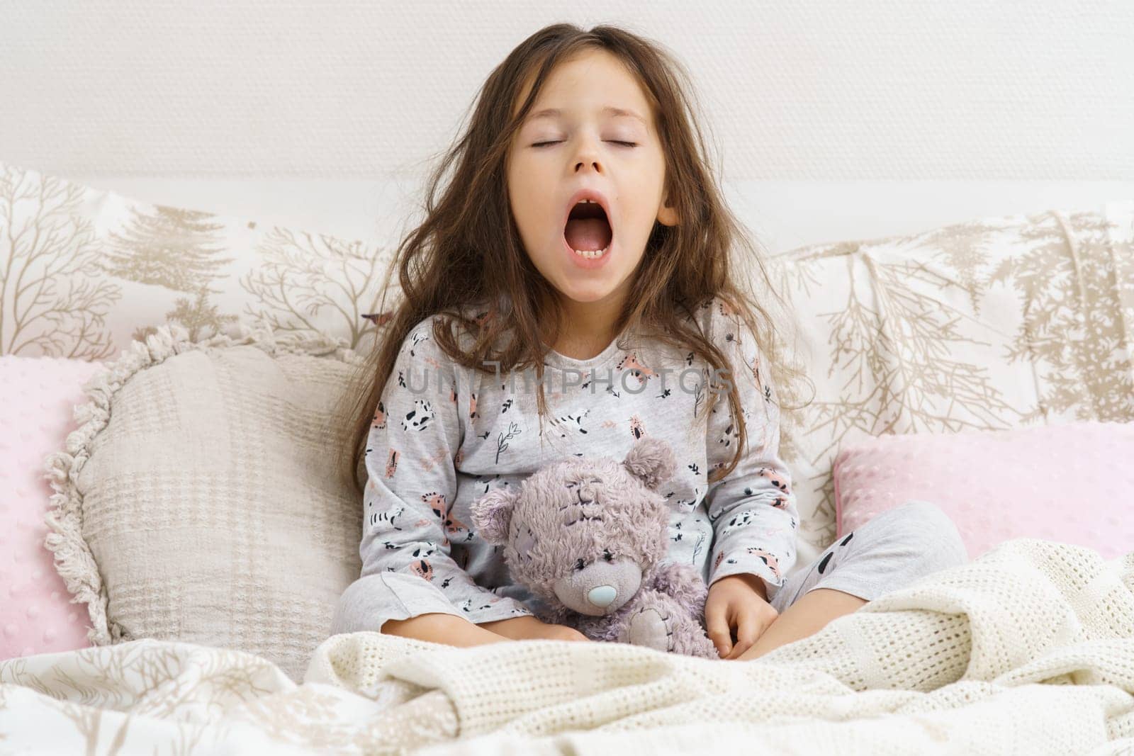 Funny, cute, tired little brunette girl yawning with open mouth, carrying teddy bear toy, sit on soft bed with pillows on white background. Broken sleep, children insomnia disorder. Copy space