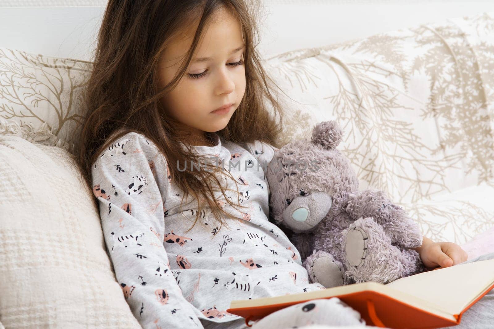 Little cute, adorable, nice brunette girl in pajamas reading book with teddy bear on soft bed. Learning at home, education and knowledge. Imagination and day dreaming. Leisure activity for introverts