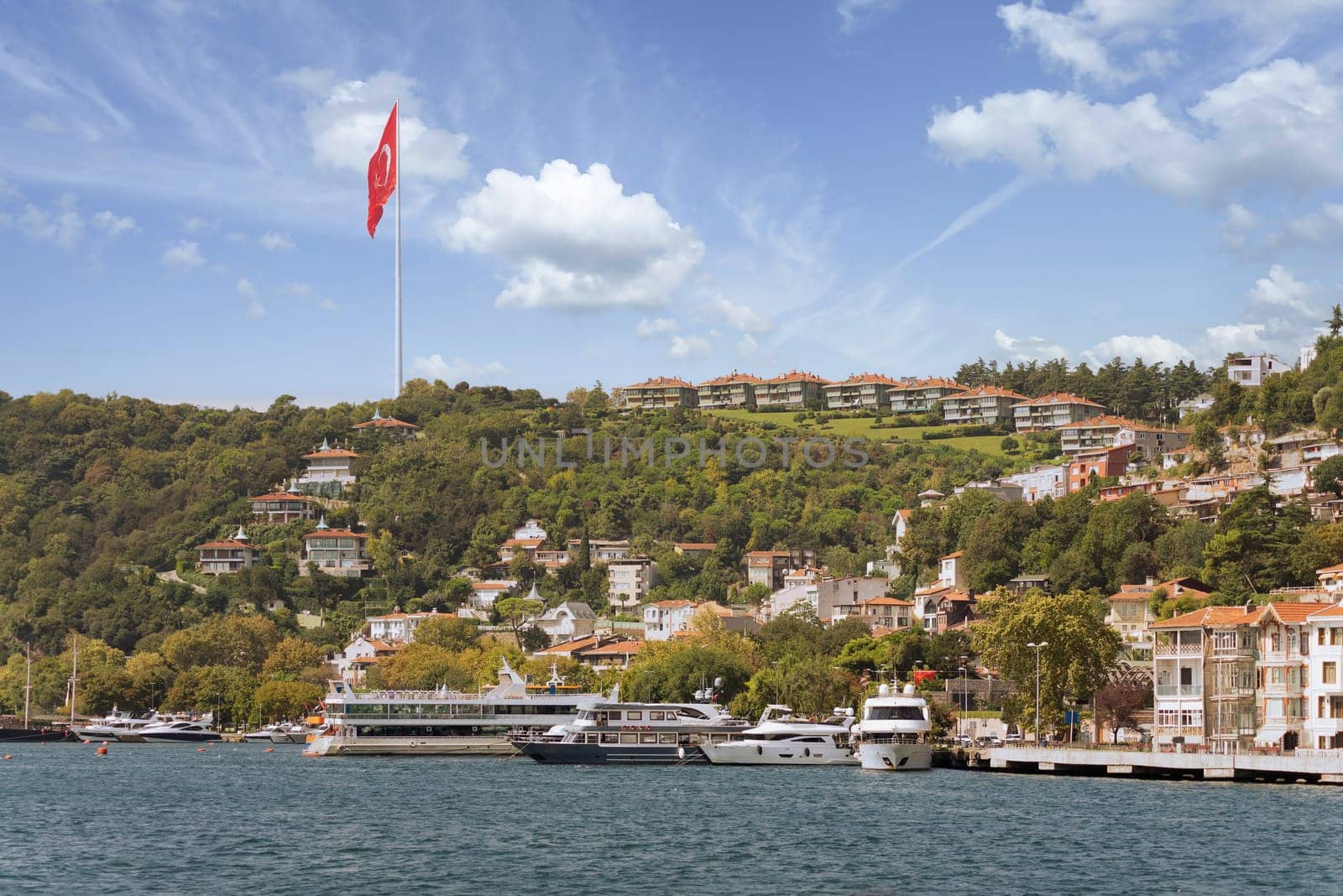 View from the sea of the green mountains of the Europian side of Bosphorus strait, with docked boats, traditional houses, dense trees, and huge Turkish flag, in a summer day