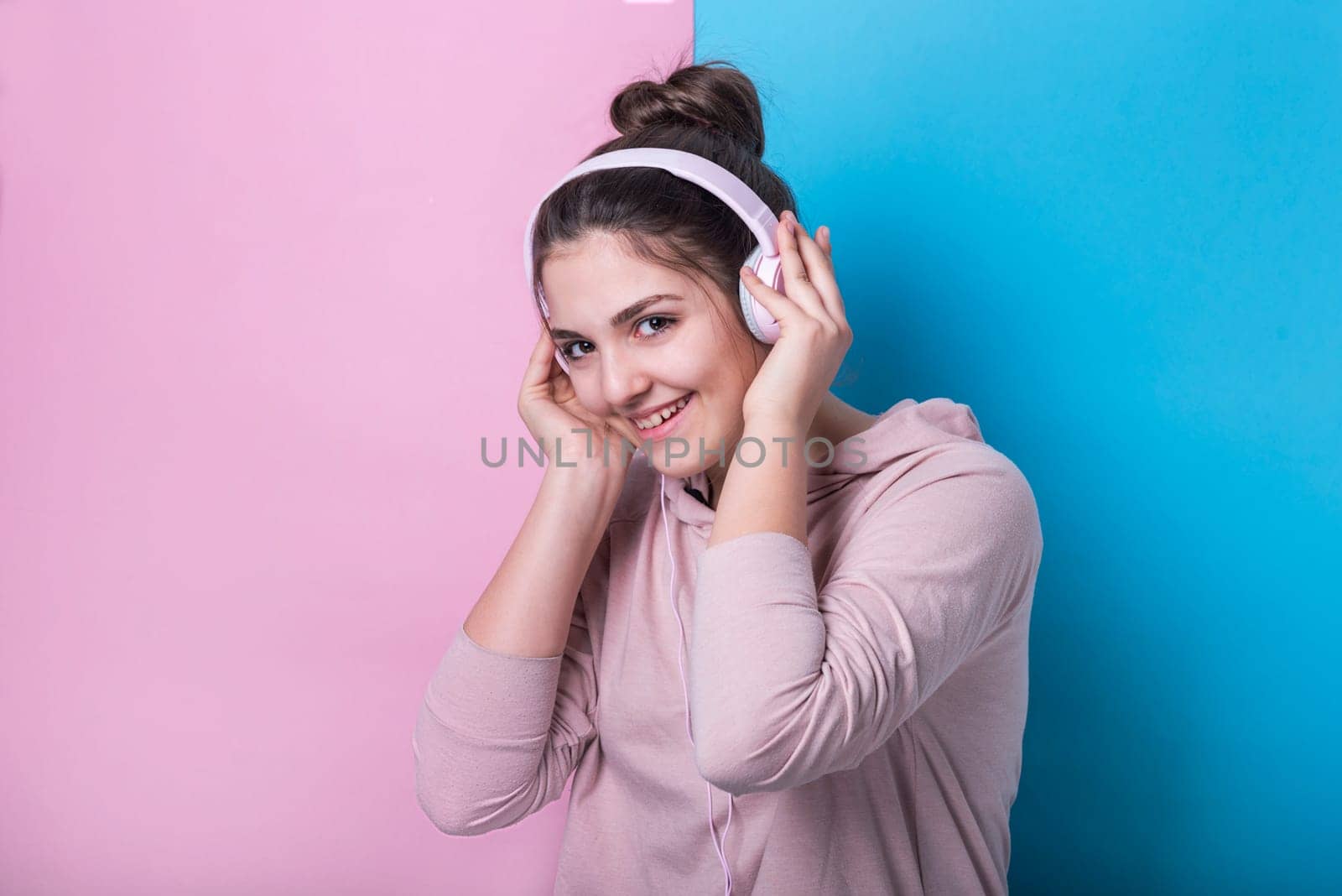 Happy teenage girl with pink headphones smiling and listening to music in front of pastel colorful studio background. Young people and youth culture lifestyle concept.