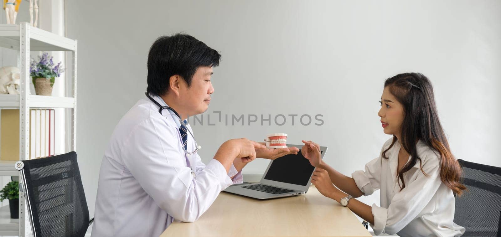 Asian dentist in white gown and stethoscope pointing finger at tooth model, explain to young patient about orthodontics and braces. Help reduce gum pain. Healthcare and medicine concept.