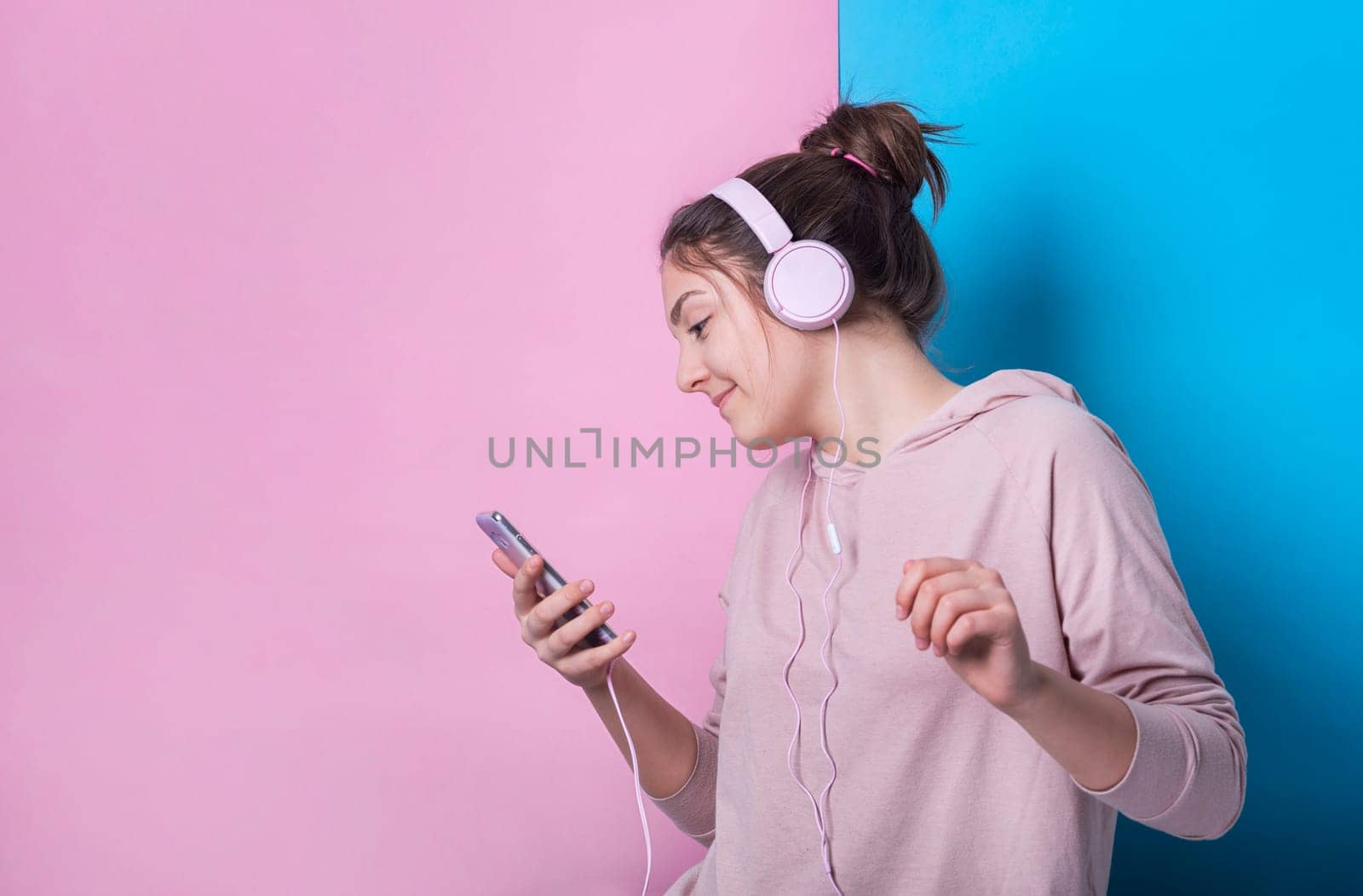 Happy teenage girl with pink headphones smiling and listening to music from her smarphone in front of pastel colorful studio background. Young people and youth culture lifestyle concept.