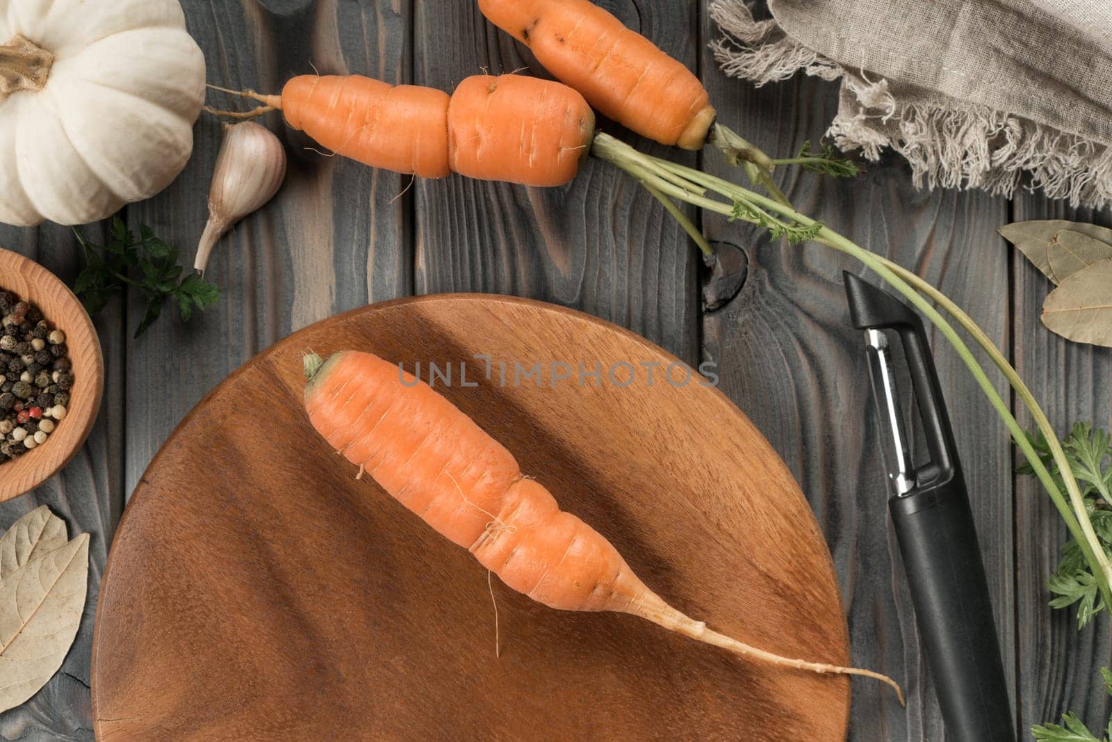 Peeling ripe orange carrot with vegetable peeler. Flat lay view. Washed unpeeled carrot. Cutting peel on kitchen wooden board. Prepare ingredients for cooking, recipe. Vegetables and flavorings.