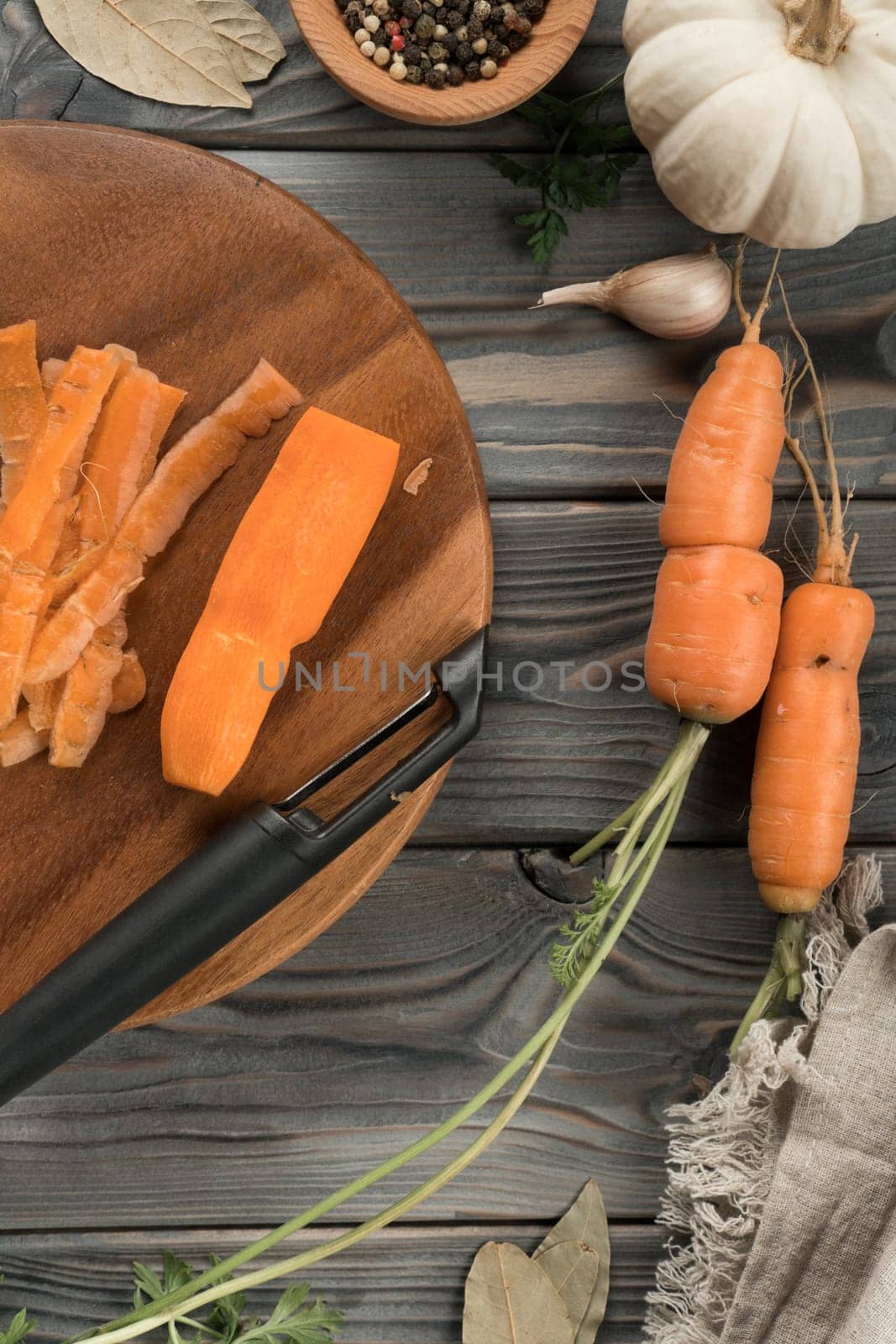 Peeling ripe orange carrot with vegetable peeler. Flat lay view. Fresh peeled carrot. Cutting peel on kitchen wooden board. Prepare ingredients for cooking, food waste. Vegetables and flavorings.