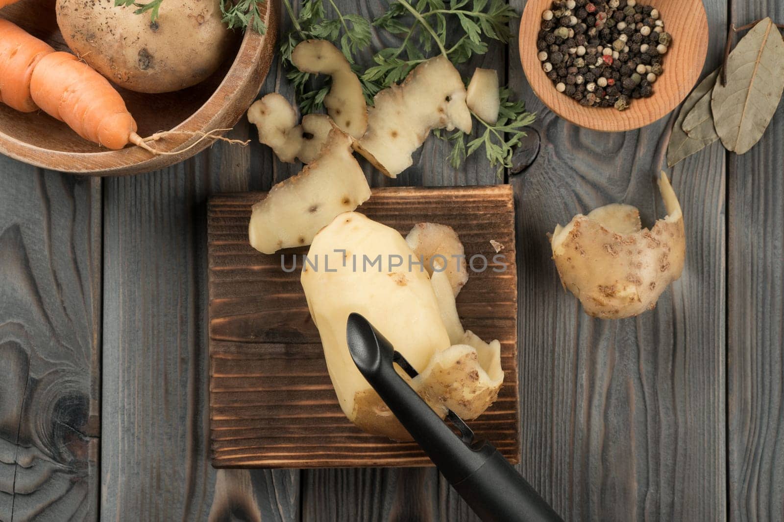 Peeling yellow potato with vegetable peeler. Flat lay view. Fresh raw potato. Skinning, cutting peel on kitchen wooden board. Prepare ingredients for cooking. Food waste. Vegetables and flavorings.