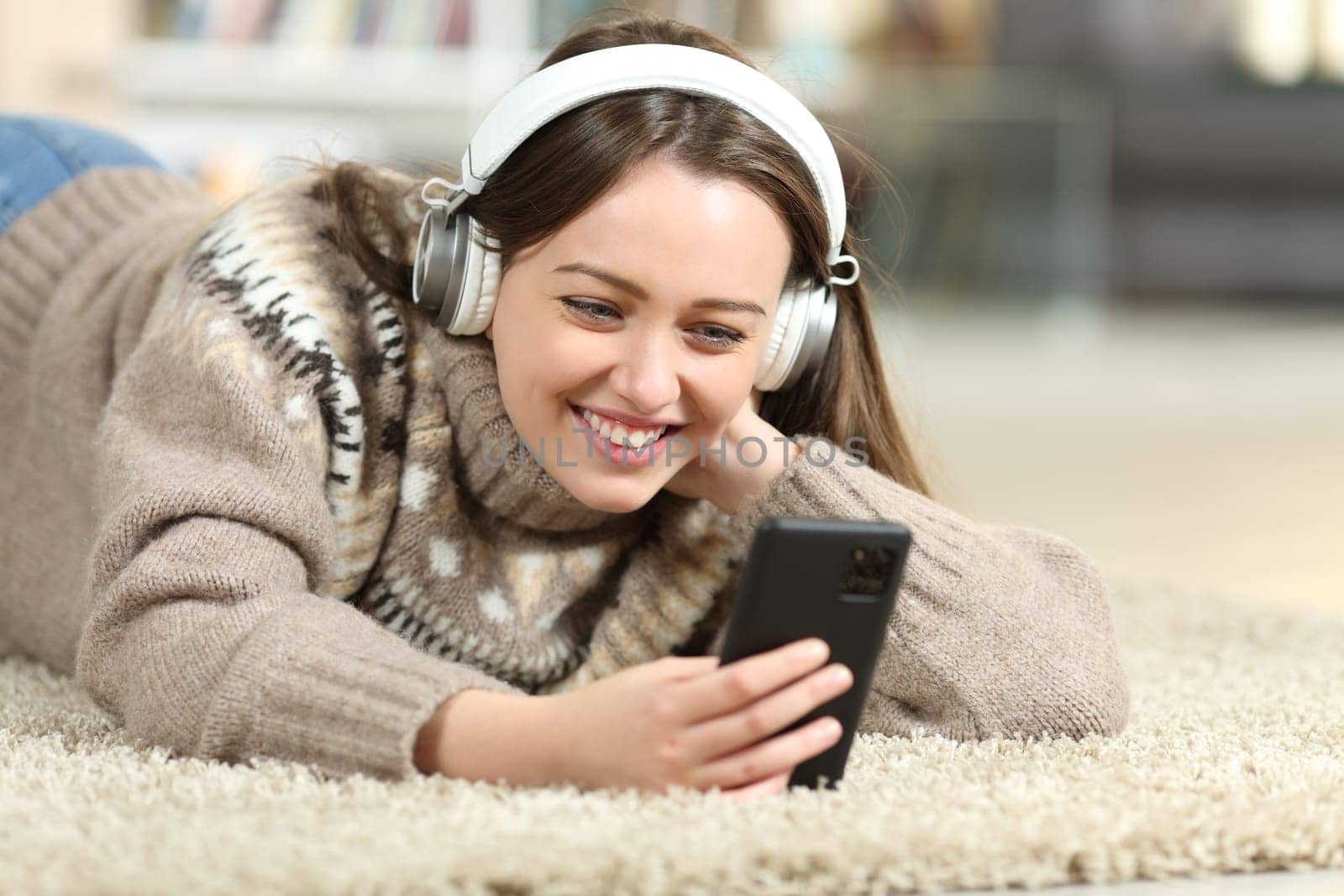 Happy teen in winter watching media on phone wearing headphones on the floor at home