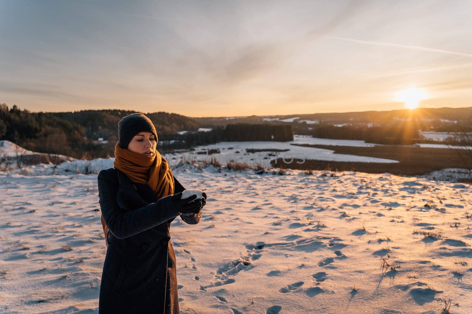 A young woman in a coat and hat at sunset stands on a snow-covered field and holds a handful of snow in her hands.