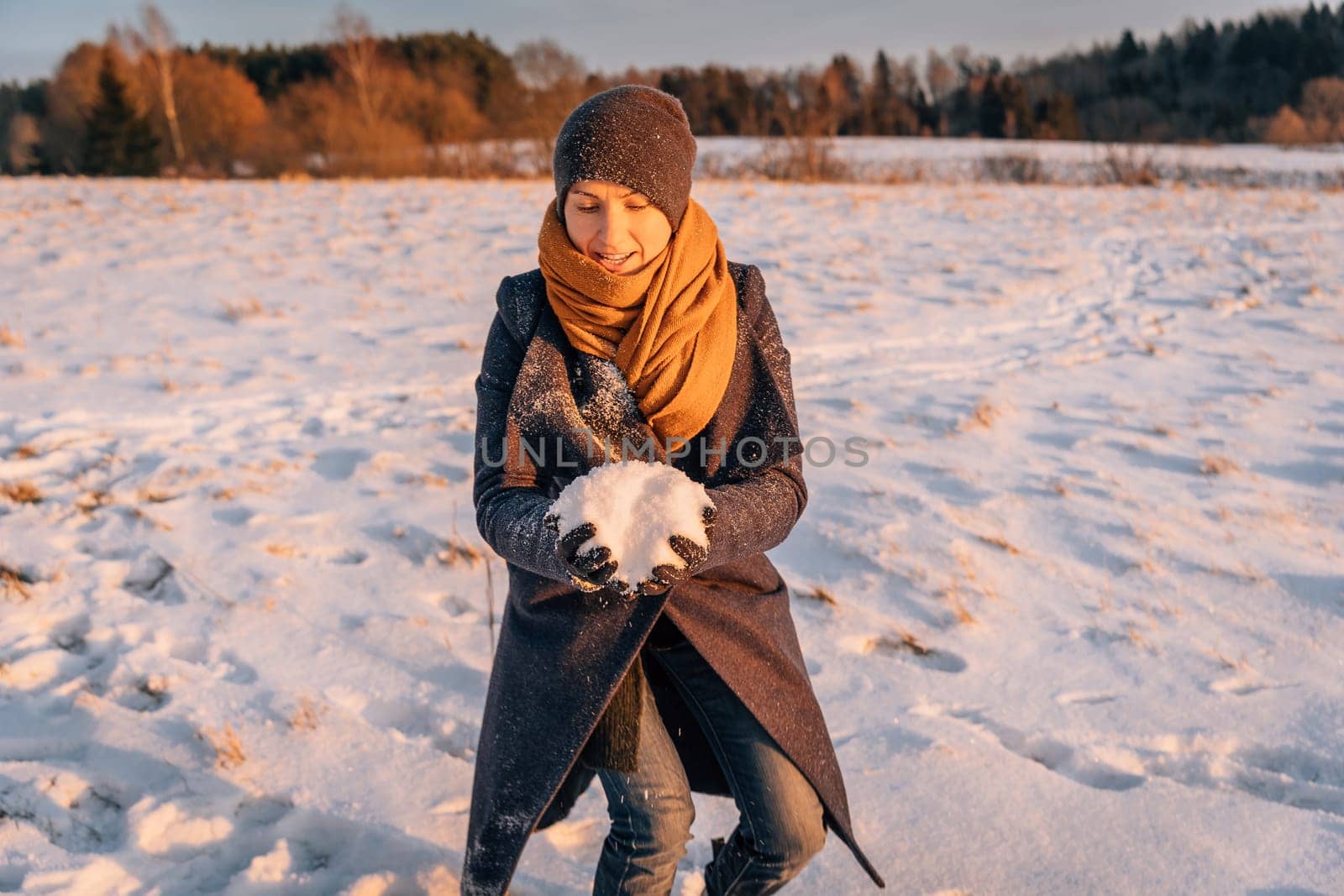 A woman in winter clothes collects a lump of snow in a snow-covered field.