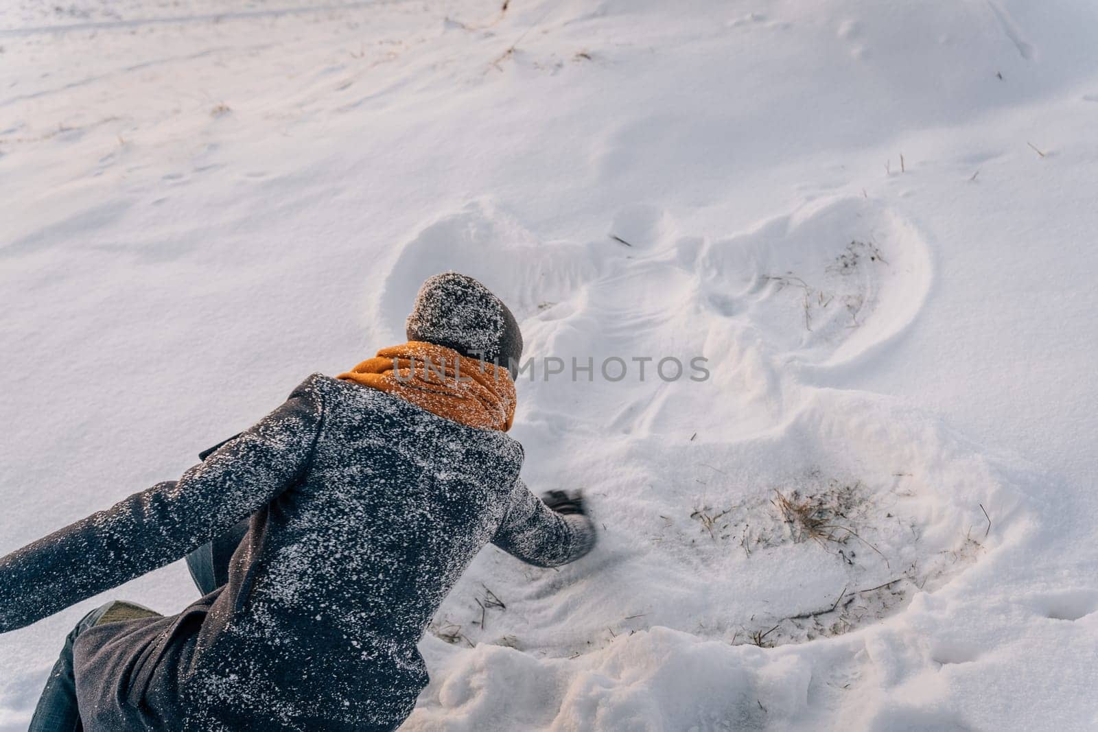A woman in a snow-covered coat and hat draws a snow angel in the snow.