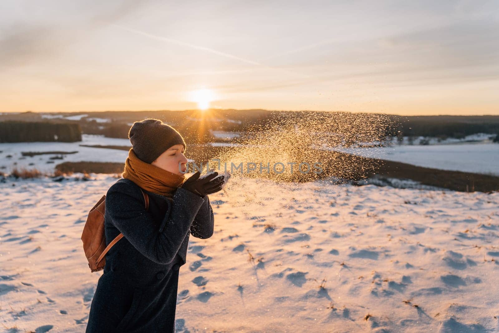 A woman on a snow-covered winter field blows snow off her palms.