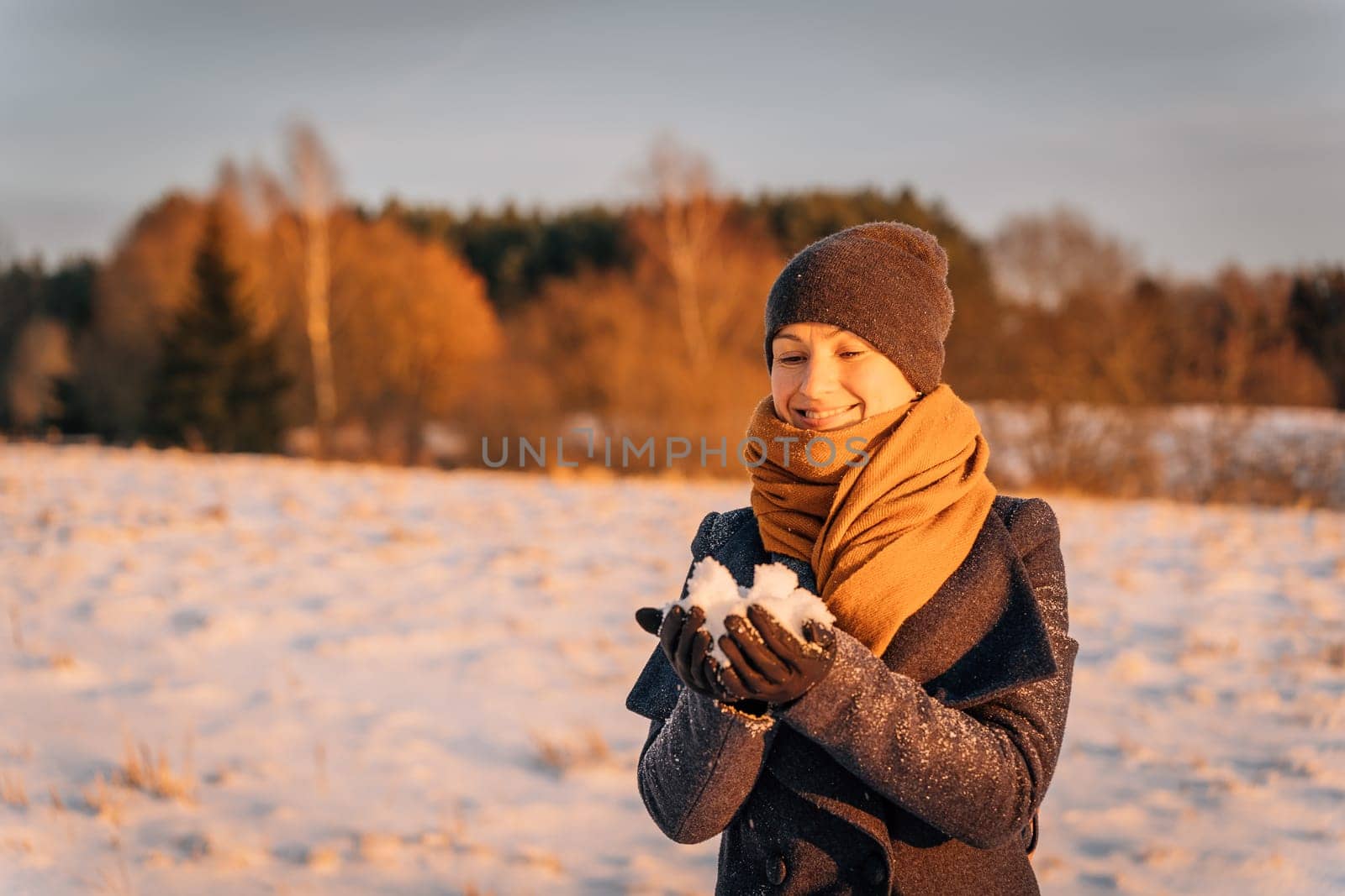 Smiling woman in a scarf and hat holds a handful of snow in winter.