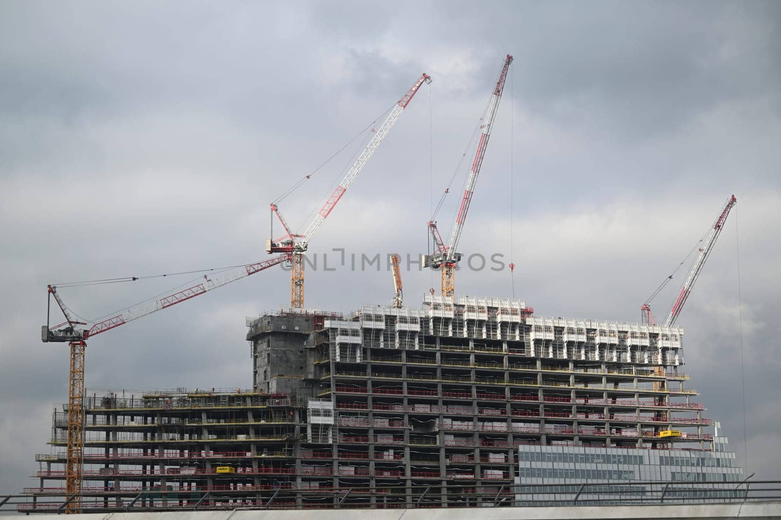 Workers are busy on a construction site in Paris, where a high-rise building is taking shape under a cloudy sky with cranes lifting materials.