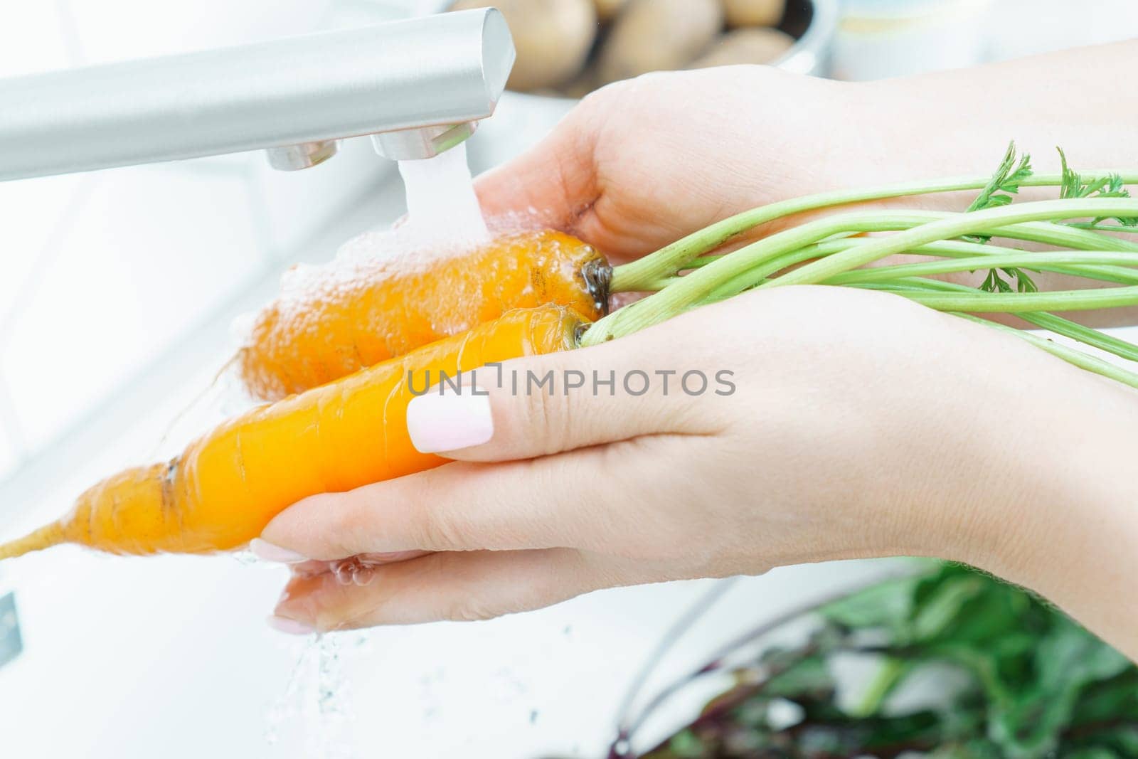 Cropped photo of woman washing orange carrots with leaves under running water from modern brass faucet in kitchen. Vegetables, vegetarian, preparation for cooking, healthy food, hygiene, vitamins.