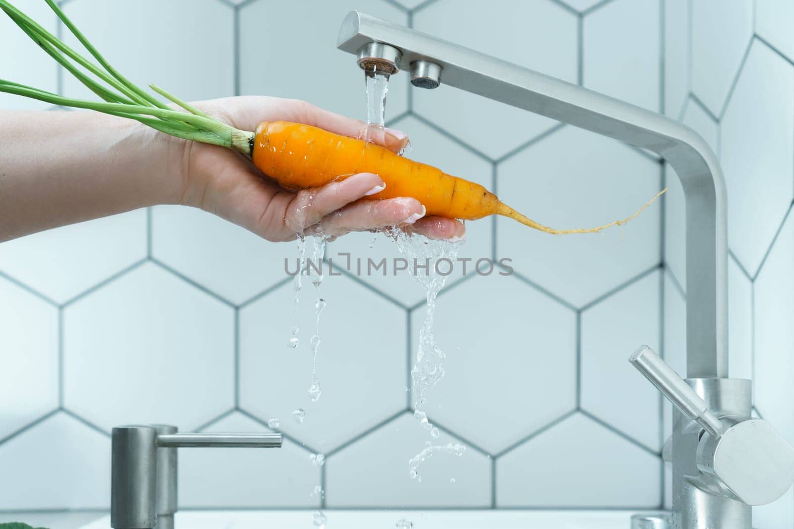 Side view of unrecognizable woman washing orange carrot with leaves under running water from modern brass faucet in kitchen. Vegetables, vegetarian, preparation for cooking, healthy food, hygiene.