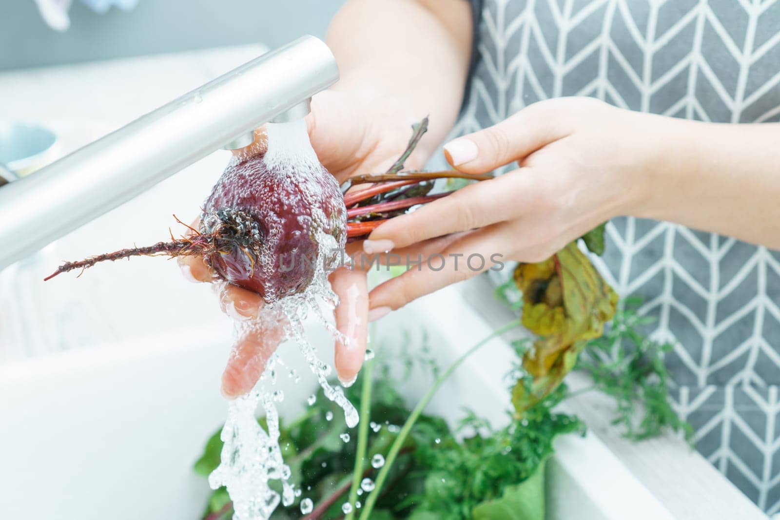 Unrecognizable woman wearing grey apron washing fresh beet with hands under running splashing water in sink full of greenery in kitchen. Vegetables, preparation for cooking, healthy food, hygiene.