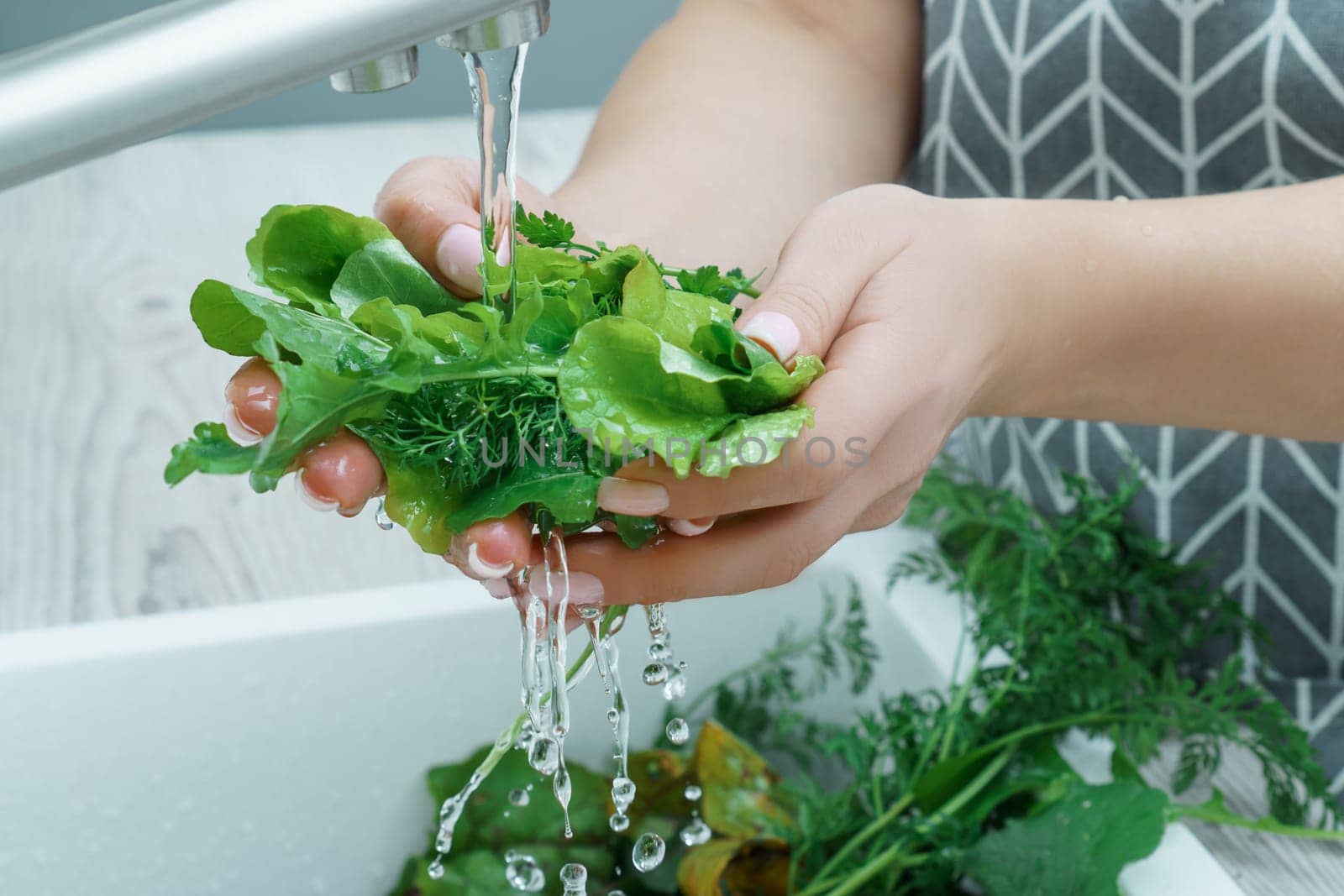 Cropped photo of woman wearing grey apron washing fresh greenery lettuce dill with hands under running water in sink in kitchen. Vegetables, vegetarian, preparation for cooking, healthy food, hygiene.