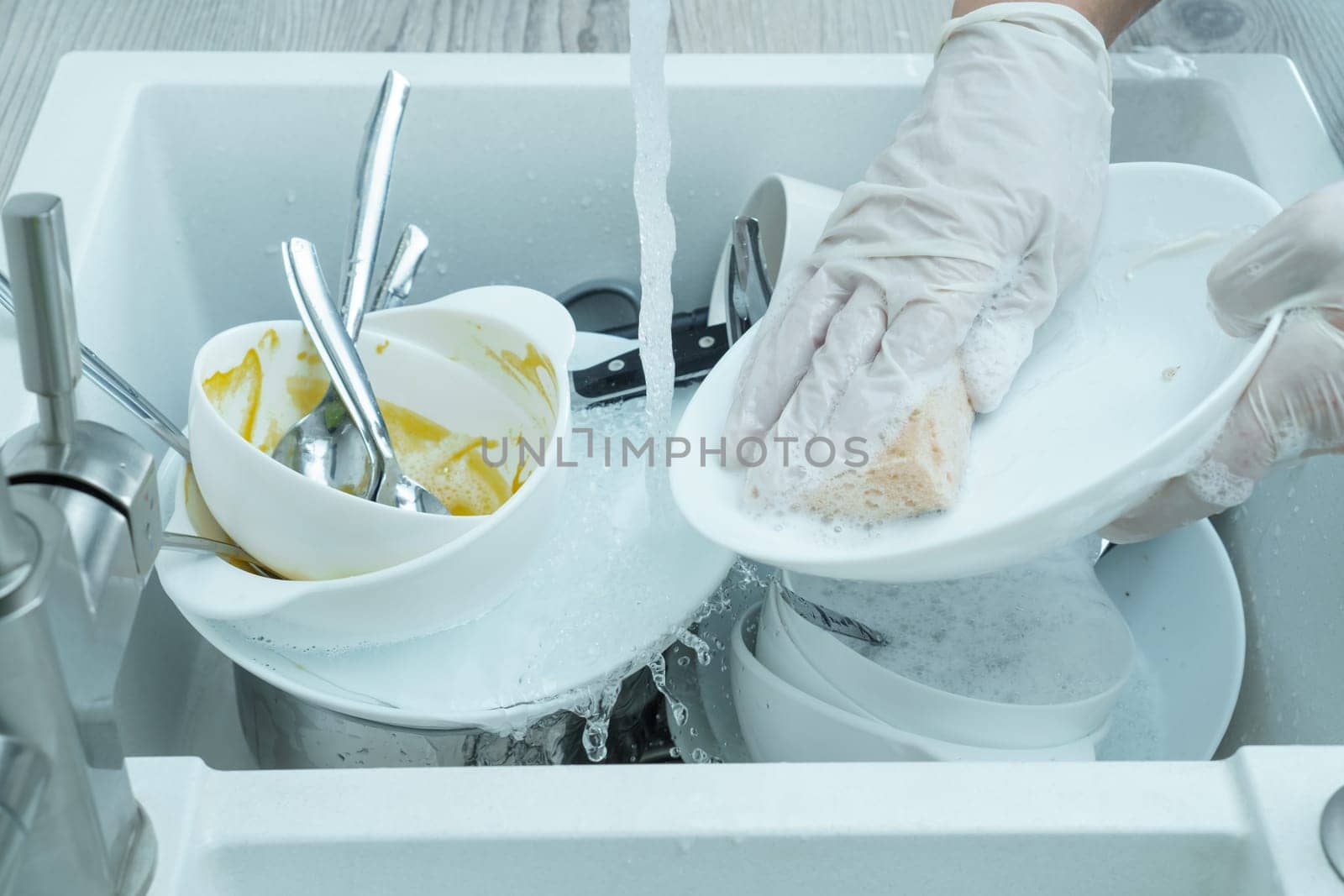 Cropped photo of woman wearing white disposable gloves, washing dirty white plate with sponge under running water in white sink full of dirty tableware. Cooking, housekeeping, cleaning, utensils.