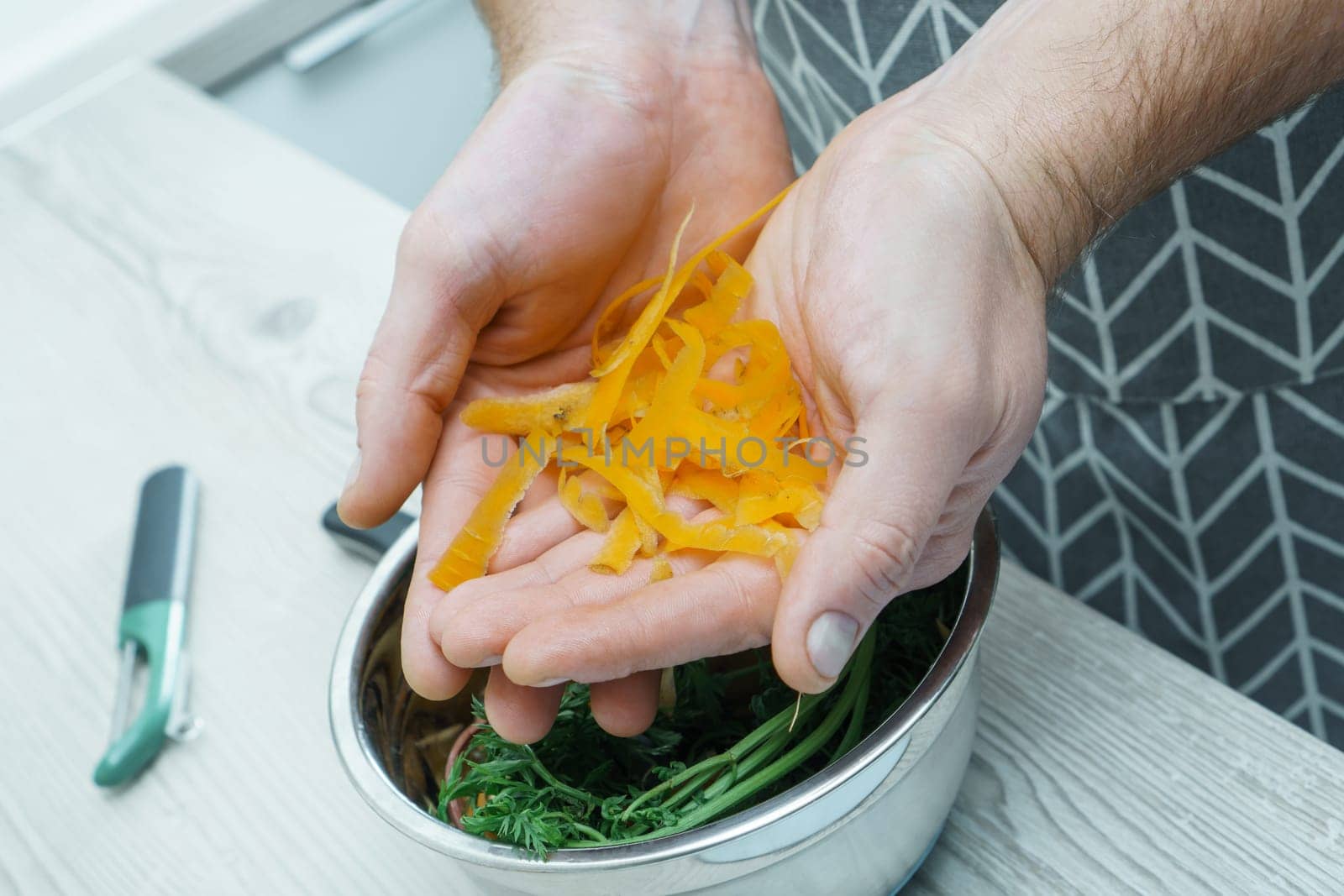 Cropped photo of man wearing grey apron holding carrot peelings in hands over metal bowl full of peelings near peeler, standing at grey wooden table in kitchen. Recycling, composting, fertilizing.