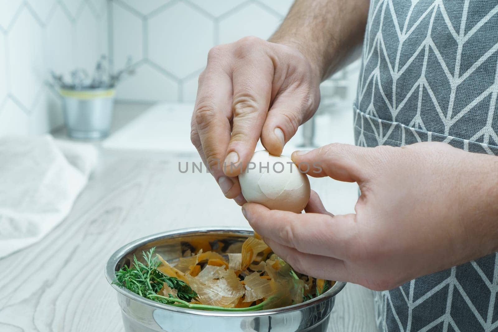Cropped photo of man wearing grey apron peeling hardboiled egg over metal bowl full of carrot, onion peelings, standing at grey wooden table in kitchen. Recycling, composting, fertilizing, zero waste.